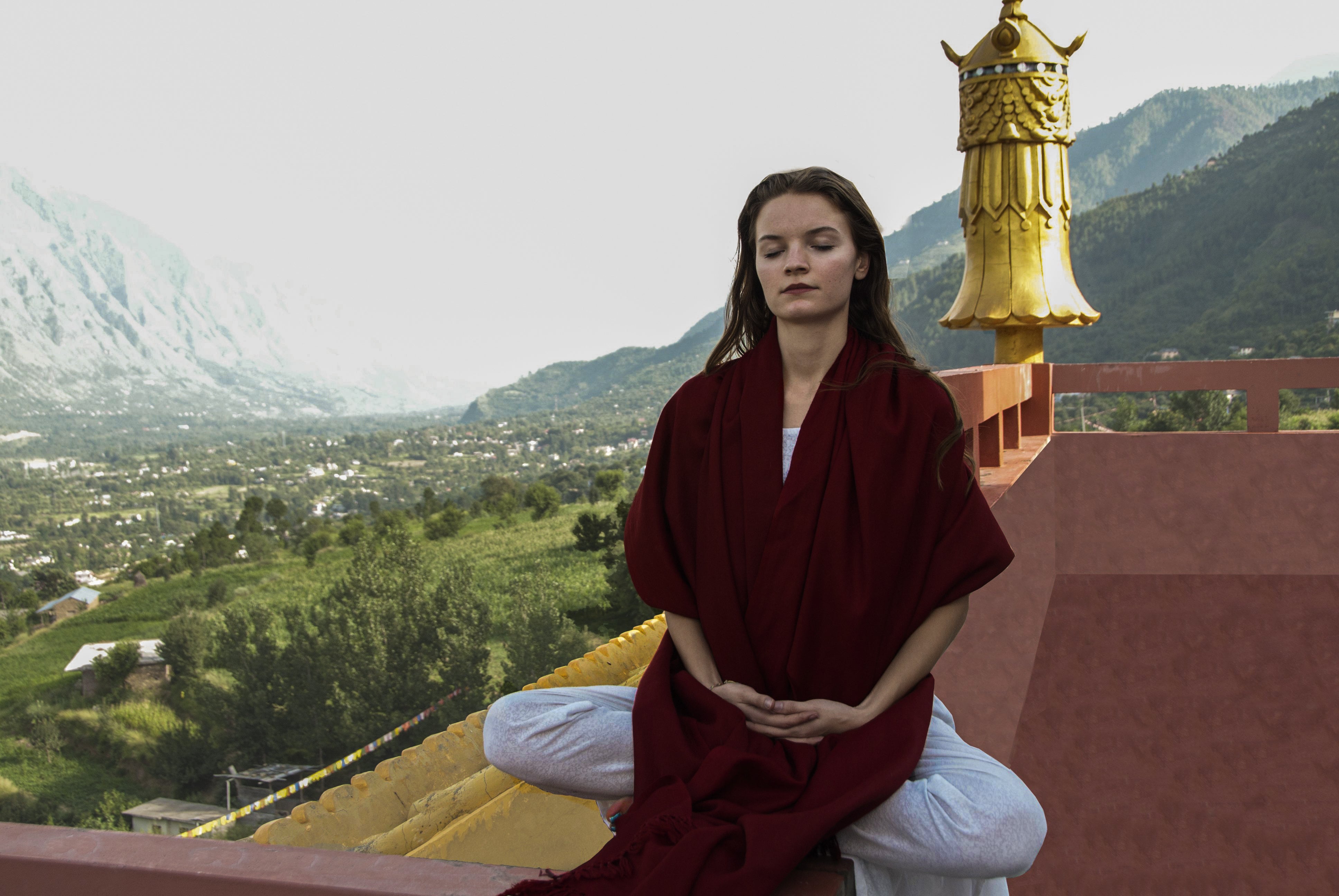 Woman over a rooftop, draped in a burgundy shawl, with a golden temple ornament and lush mountain view.