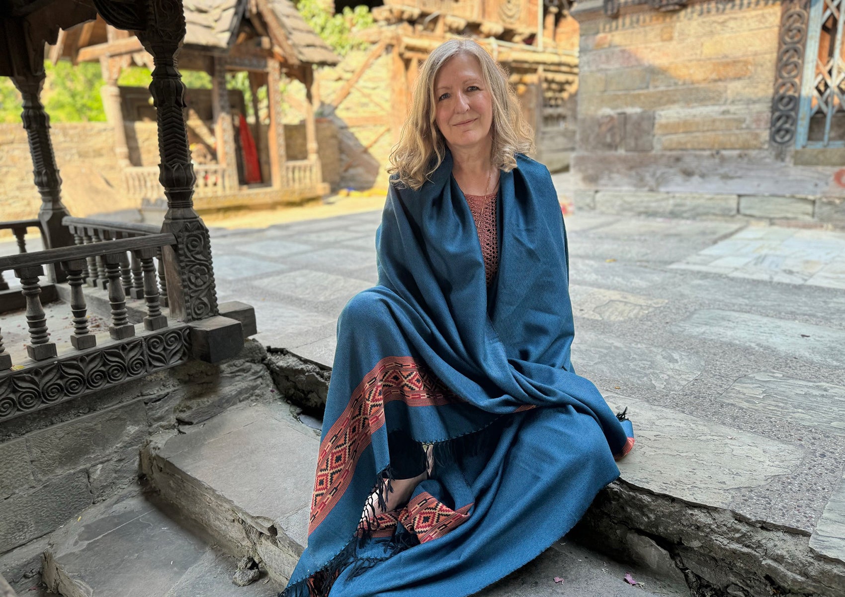 Woman sitting serenely in a stone courtyard, wrapped in a denim blue shawl with intricate red detailing