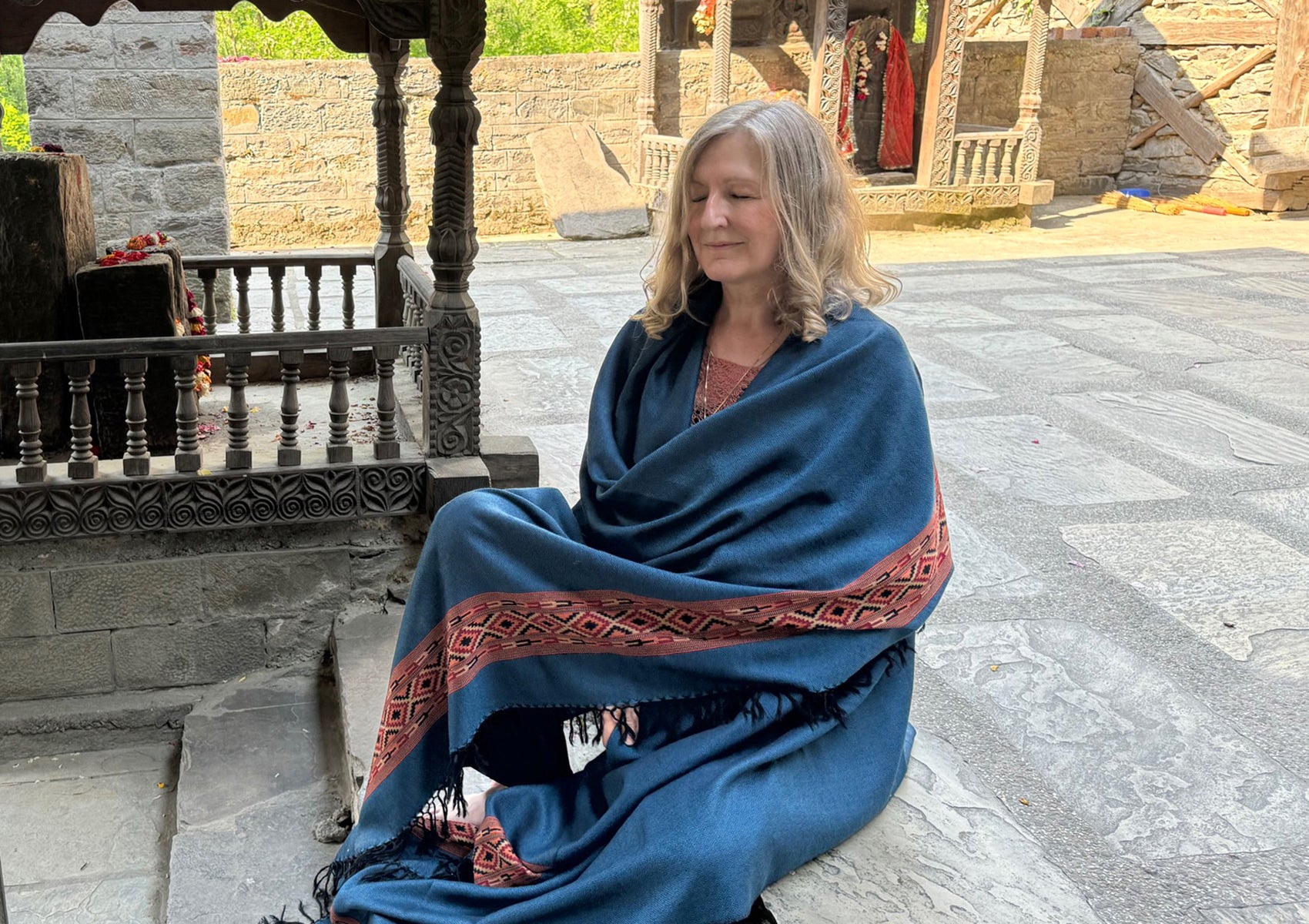 Woman in a meditative pose wrapped in a denim blue shawl with intricate red and black embroidery detailing, seated near traditional carved stone structure