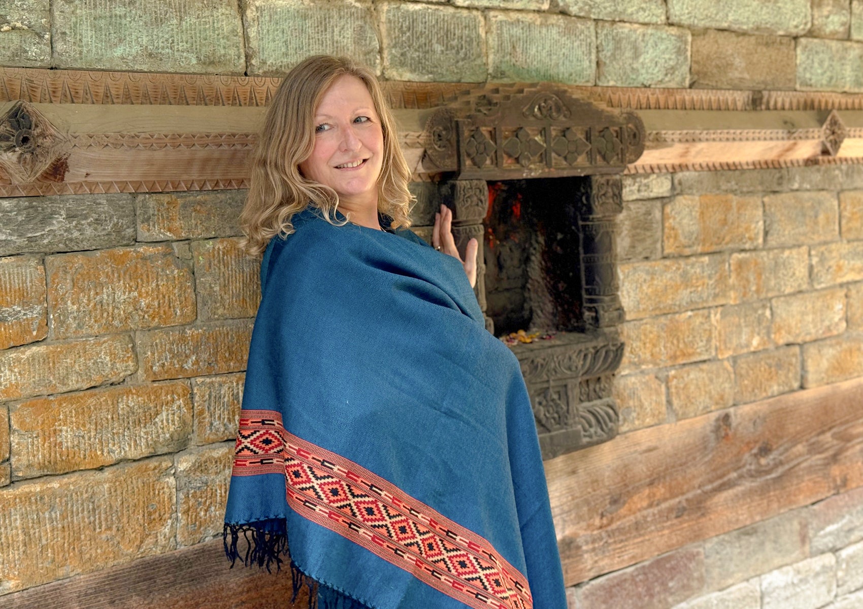 Smiling woman draped in a denim blue shawl with red patterned trim, standing beside a carved stone wall