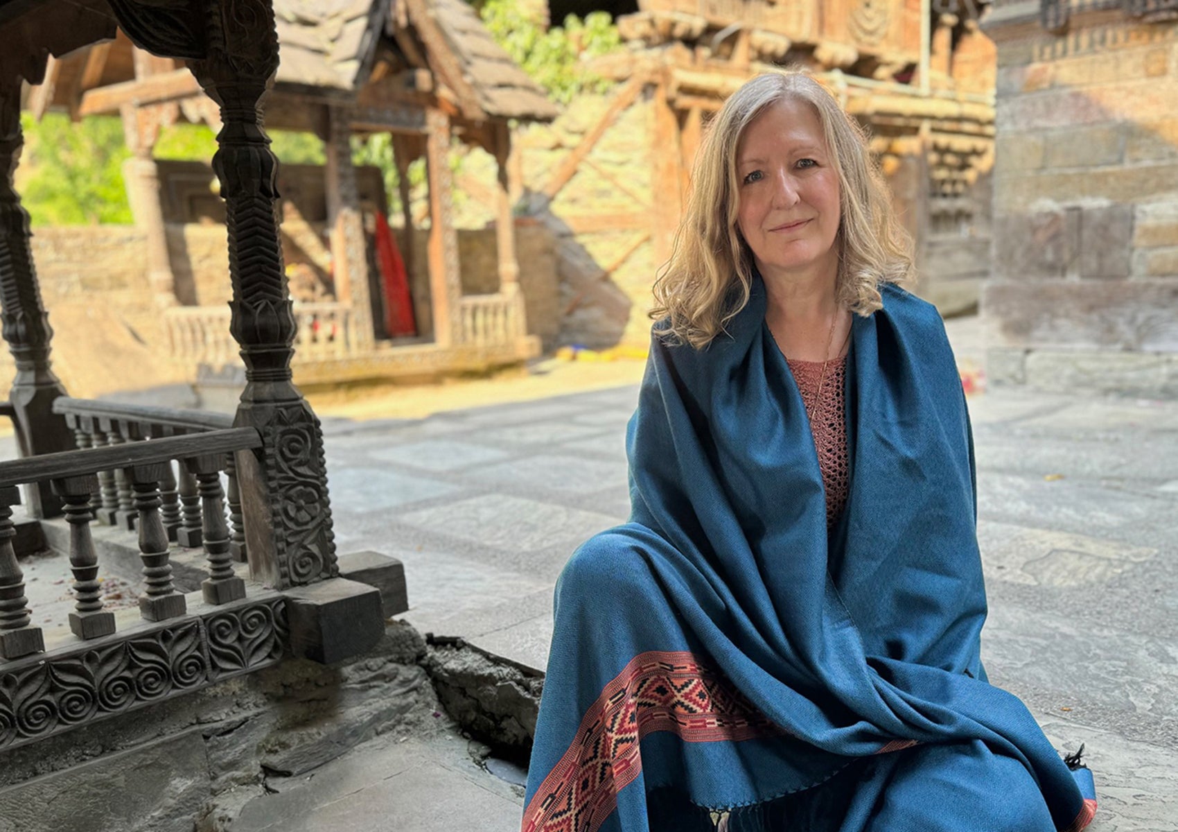 Woman sitting near ornate temple carvings, wrapped in a denim blue shawl with intricate red and black embroidered patterns