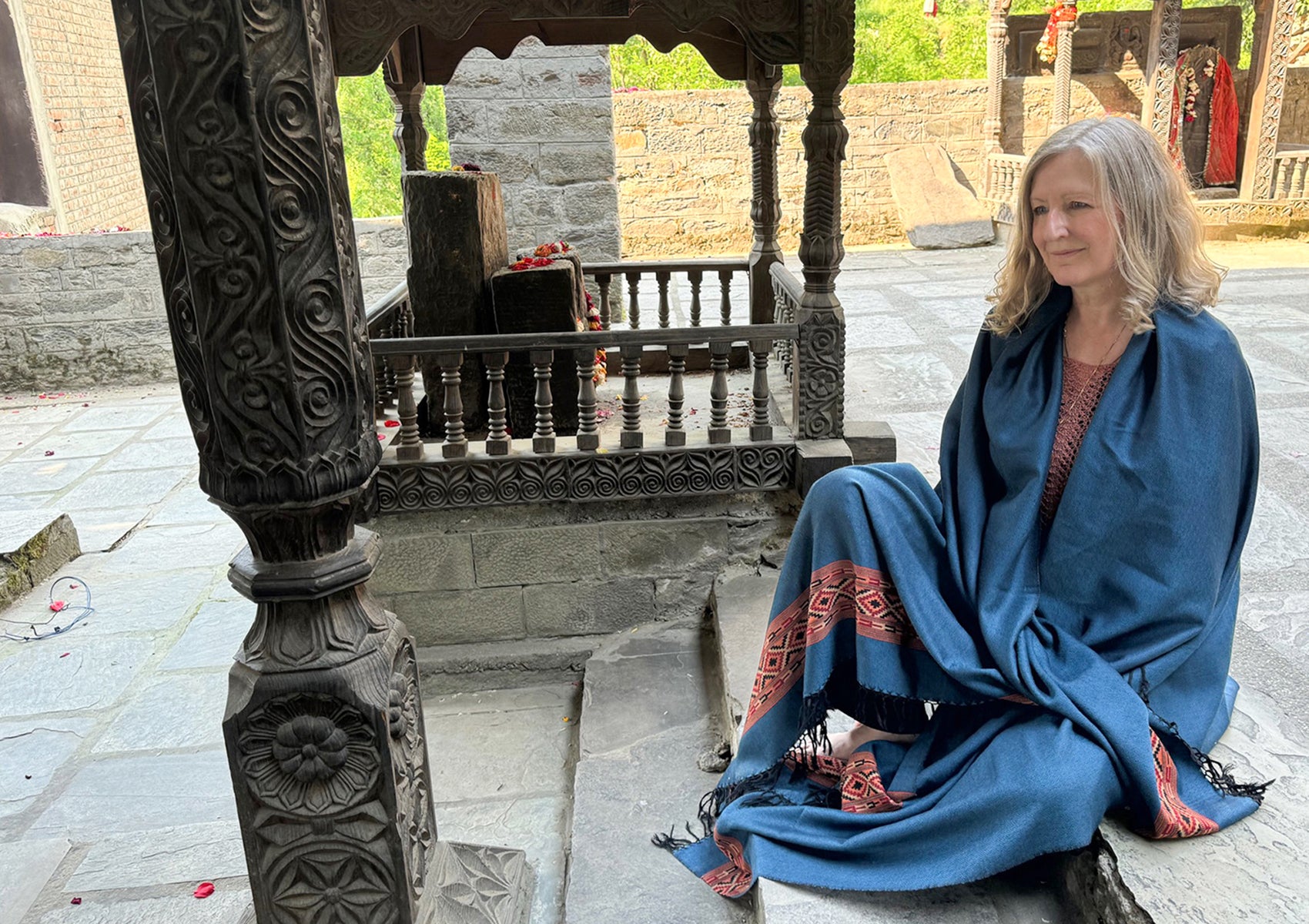 Woman sitting near an intricately carved stone structure, wrapped in a denim blue shawl with red and black geometric embroidery