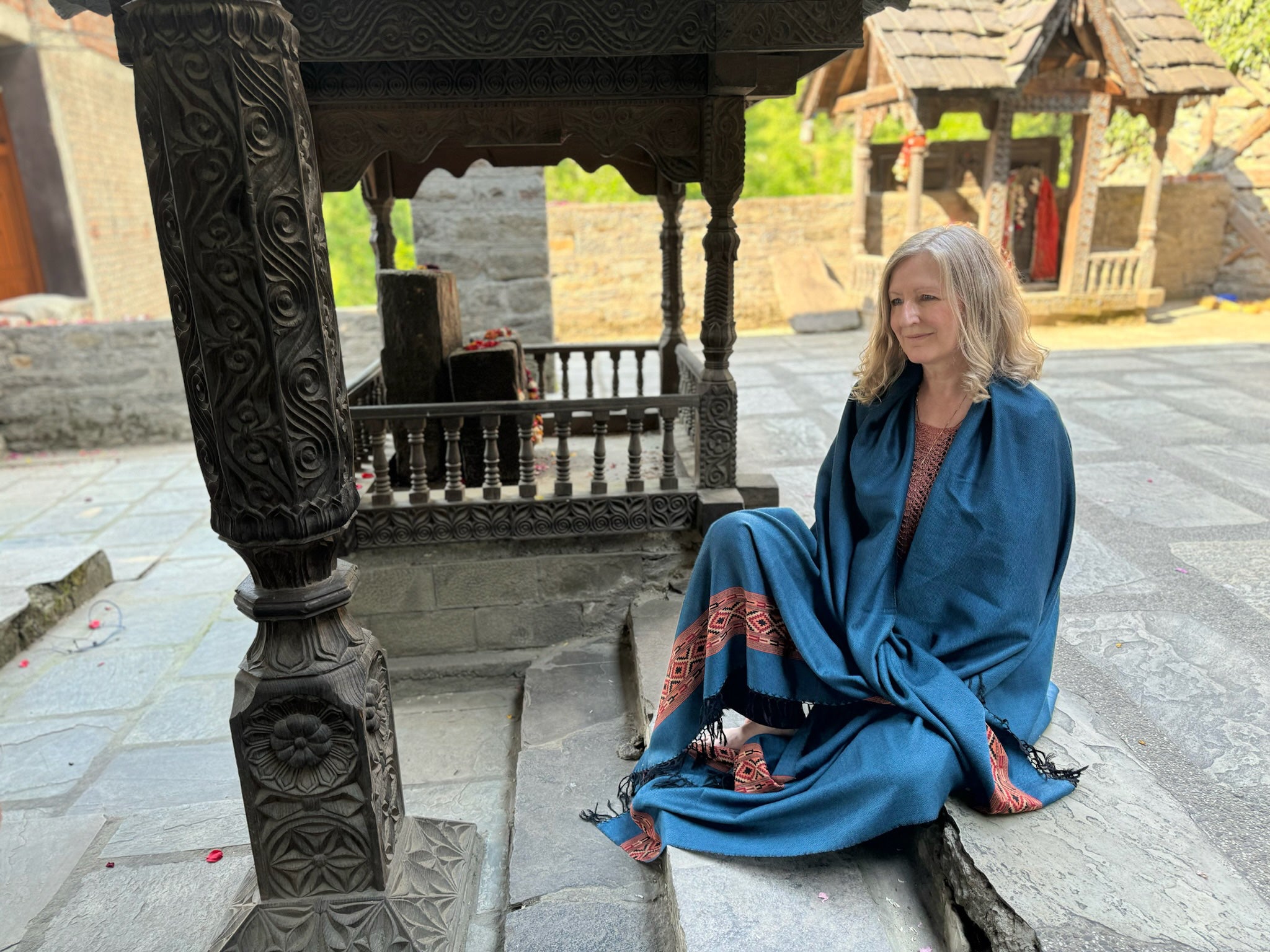 Woman sitting peacefully by a carved stone pillar in a temple courtyard, wrapped in a denim blue shawl with red woven detailing