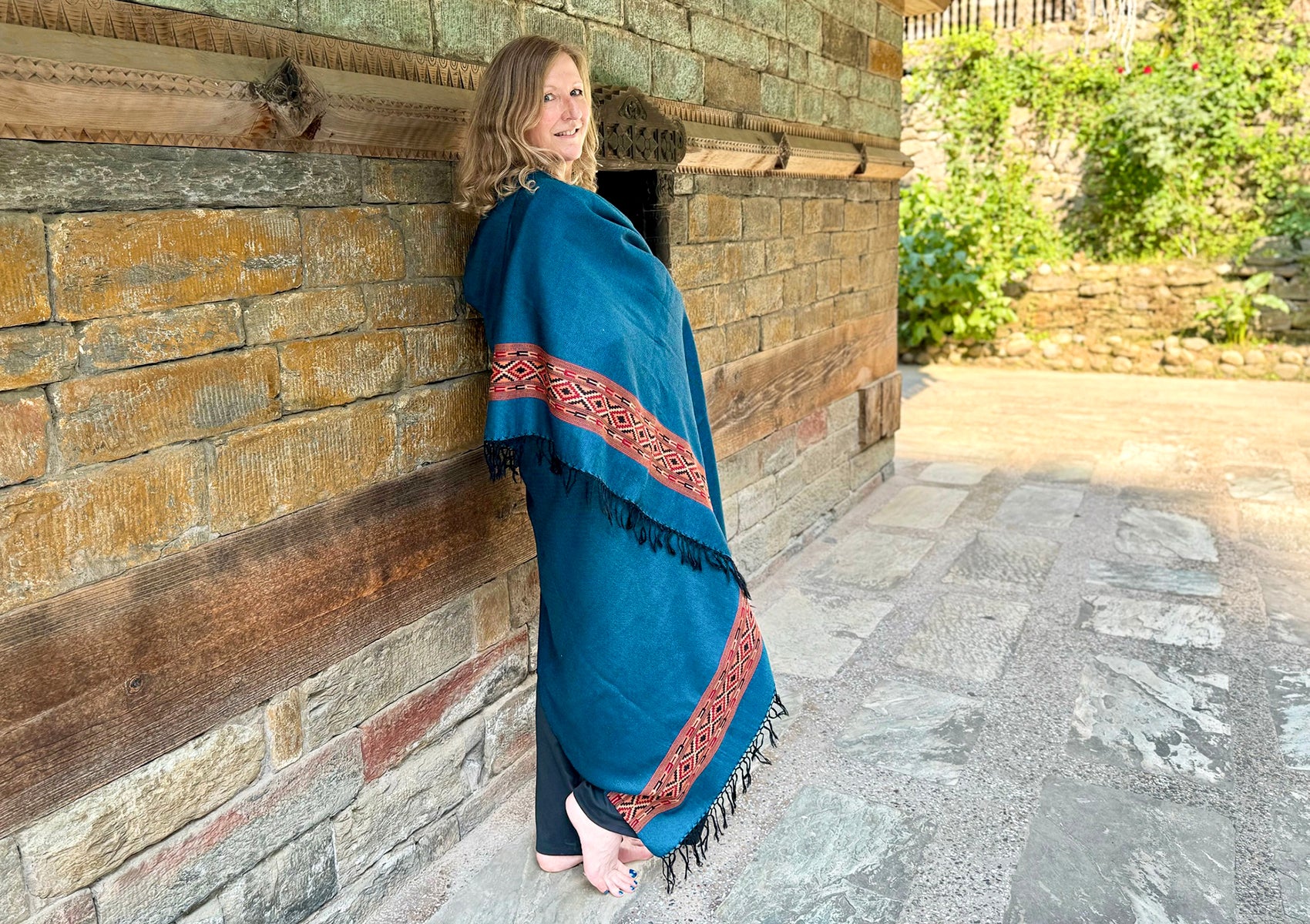 Woman standing against a rustic stone wall, wrapped in a denim blue shawl with red and black intricate woven accents