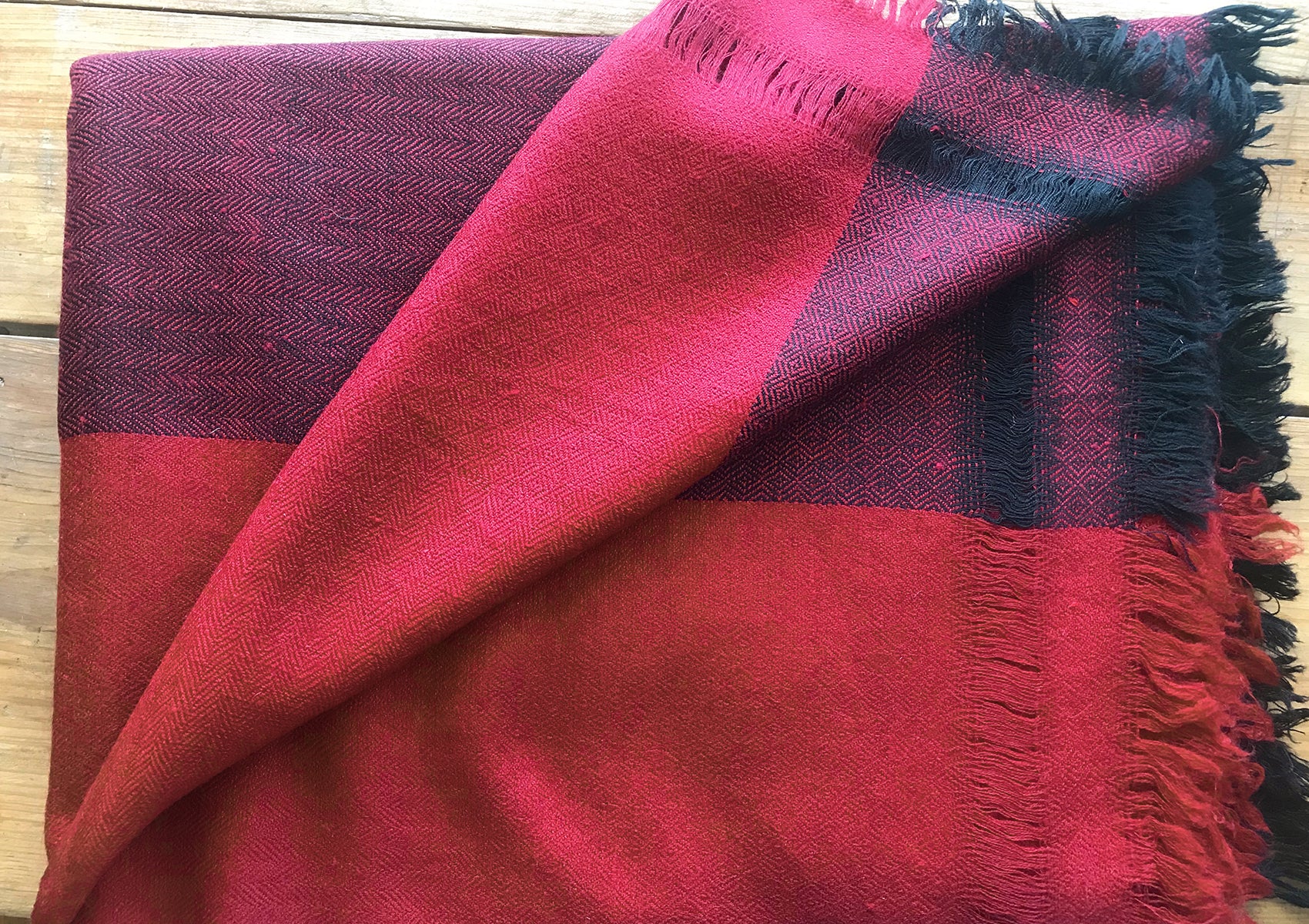 Close-up of a folded burgundy shawl with black fringe, showcasing the intricate woven texture and rich color contrast on a wooden surface.