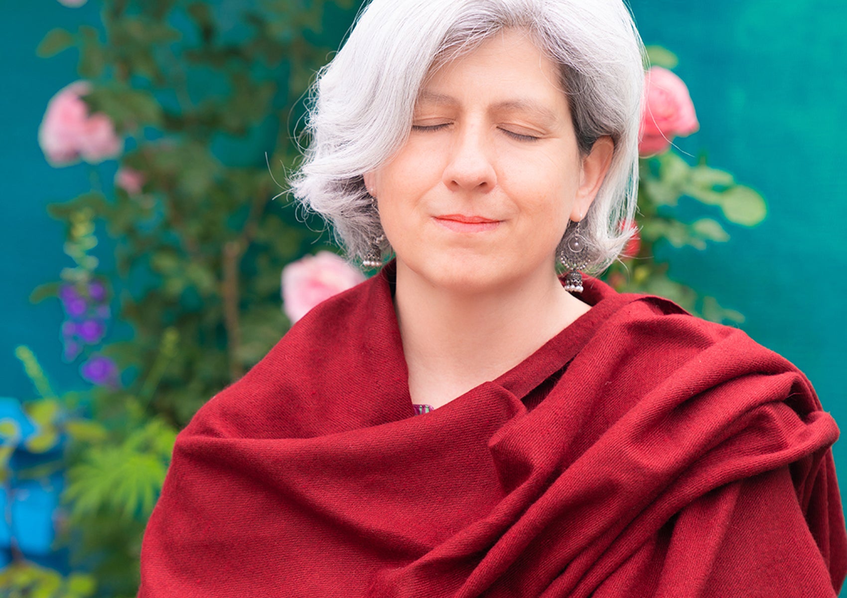 Woman with gray hair draped in a handmade burgundy shawl, eyes closed in a peaceful expression, standing in a vibrant garden with flowers.