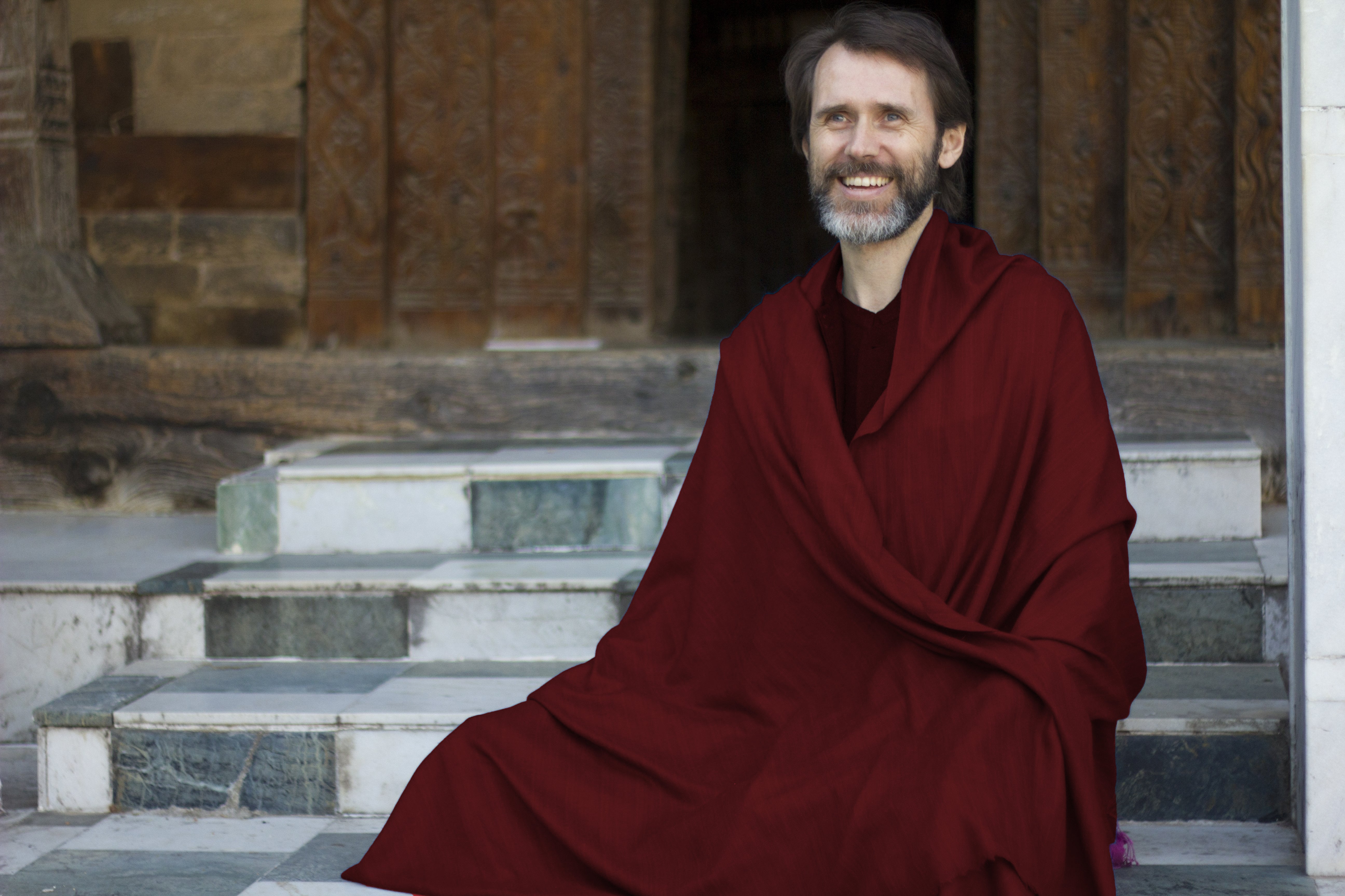 A man seated on marble steps, wrapped in a red Pashmina meditation shawl, with an intricately carved wooden temple backdrop, exuding peace and tradition.