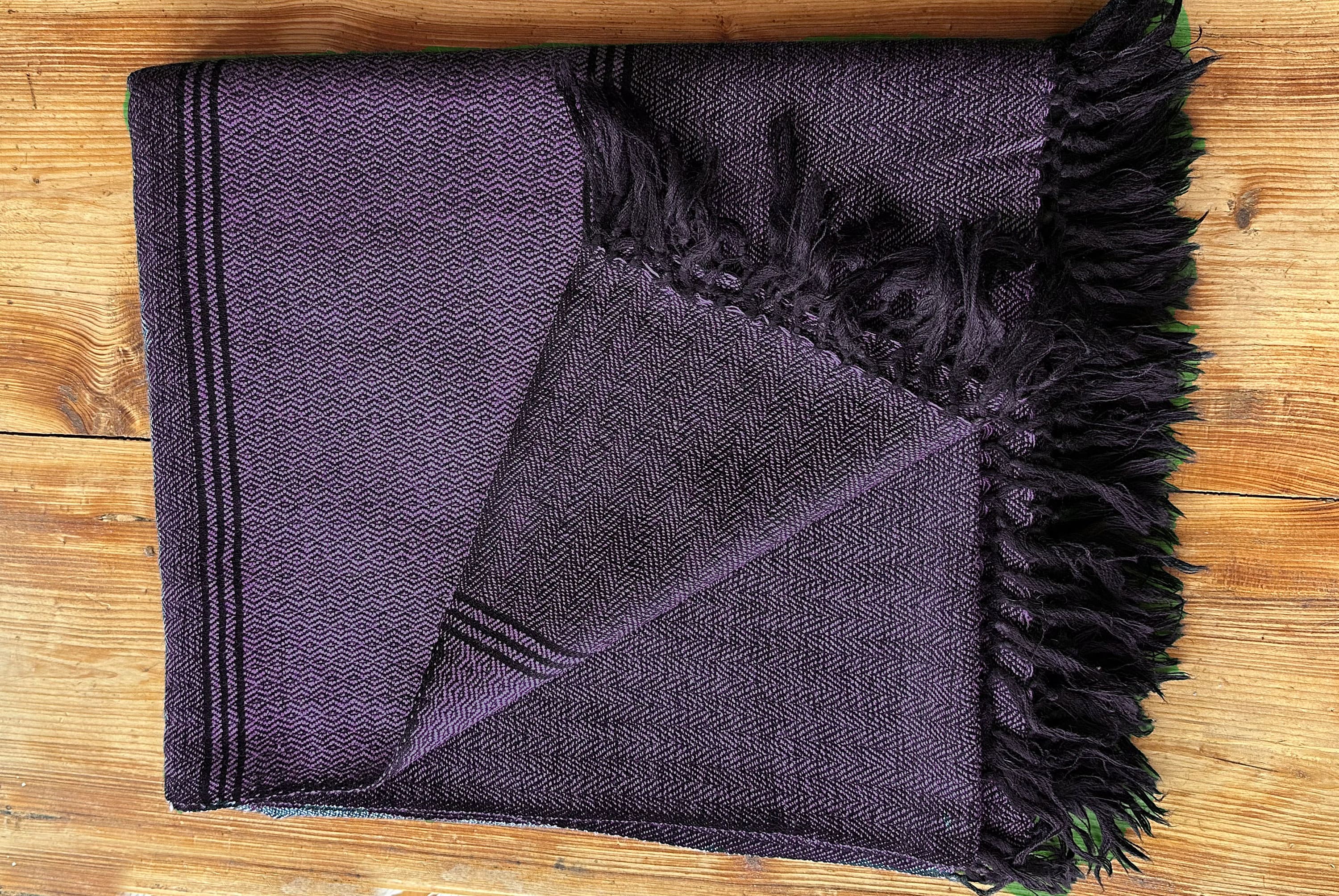 Purple Himalayan blanket neatly folded with black tassels, displayed on a rustic wooden background.