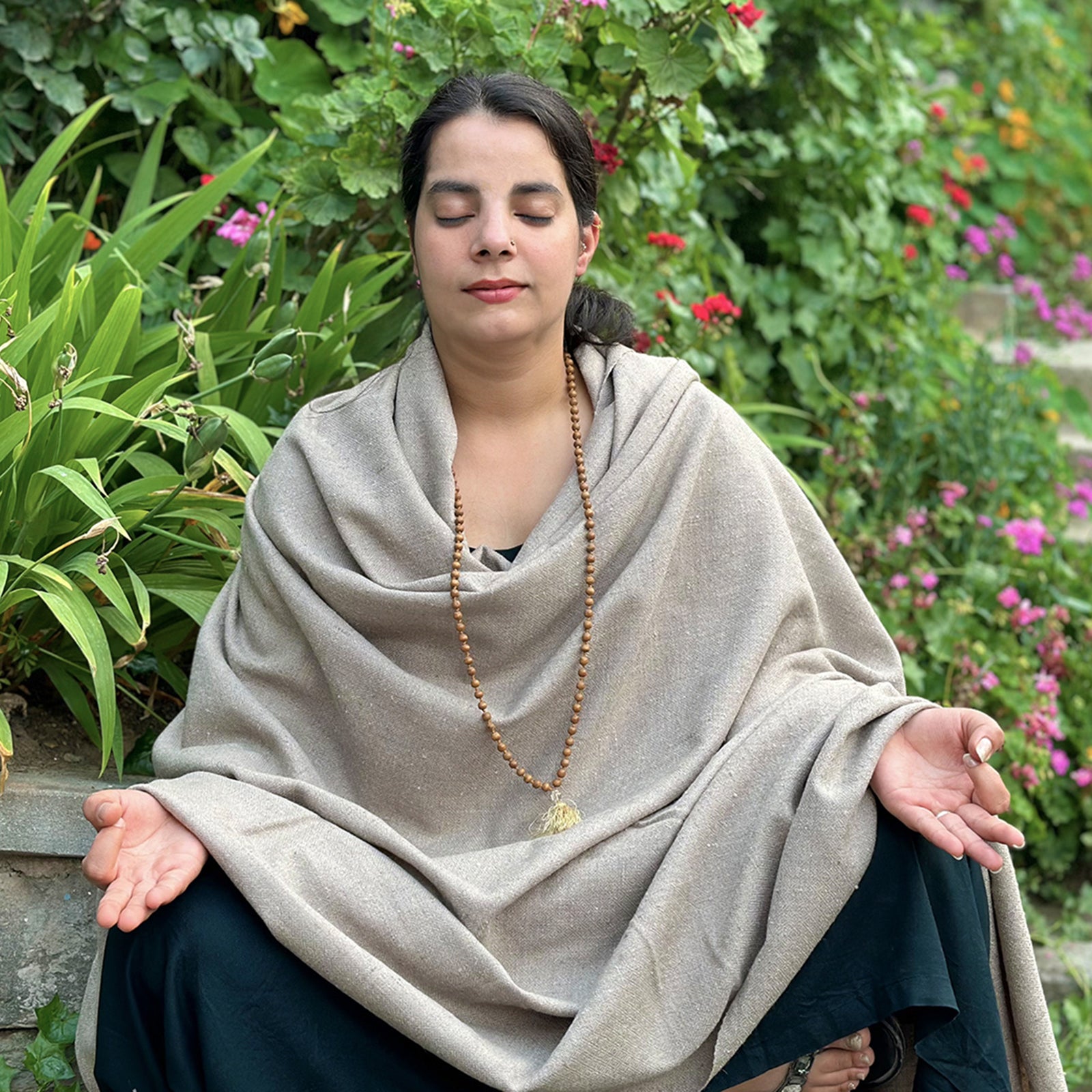 Individual sitting in meditation posture with a beige Kailash shawl, surrounded by lush green foliage and colorful flowers.