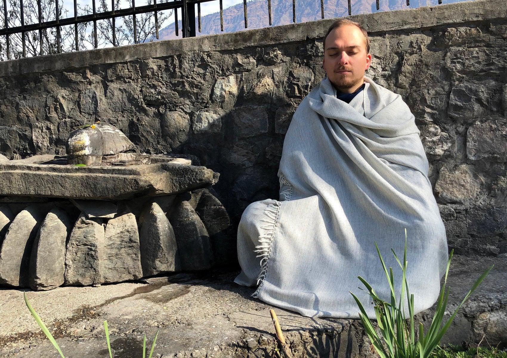 Man meditating outside, wrapped in a grey shawl, seated by a stone structure with plants around.