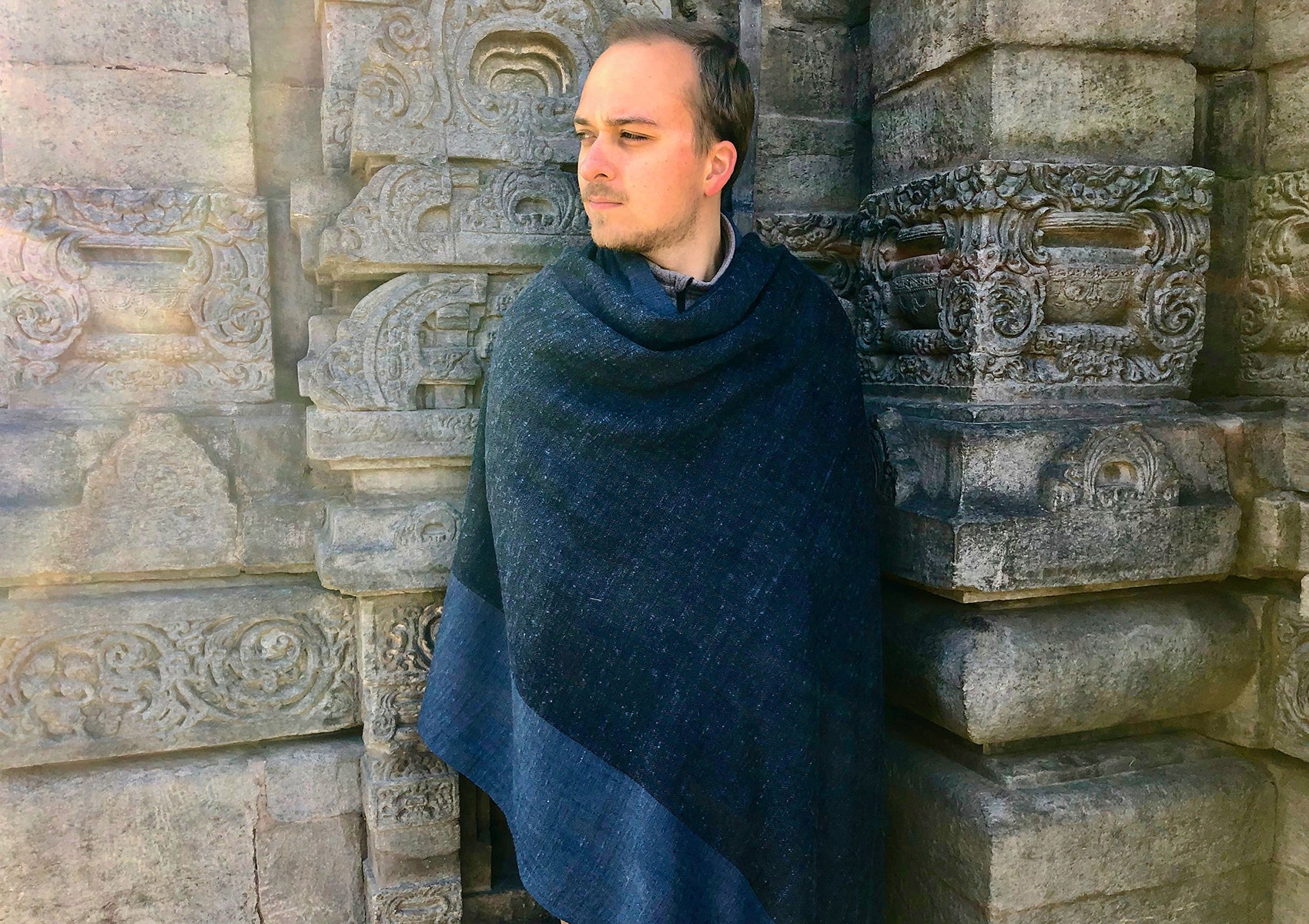 Man draped in a large blue shawl, standing pensively beside a stone wall with intricate temple carvings, merging traditional design with contemporary style.