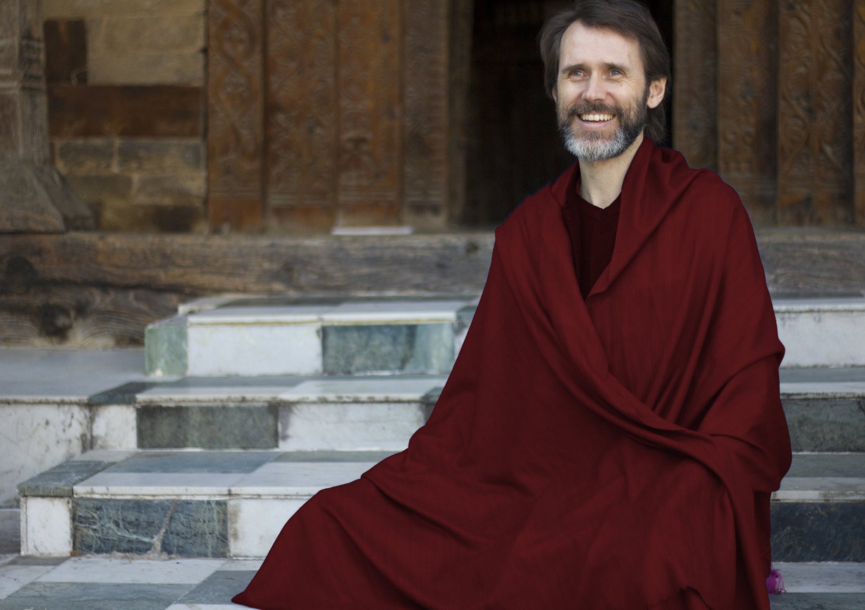 Man meditating outdoors, wrapped in a burgundy shawl, sitting on temple steps with a peaceful expression