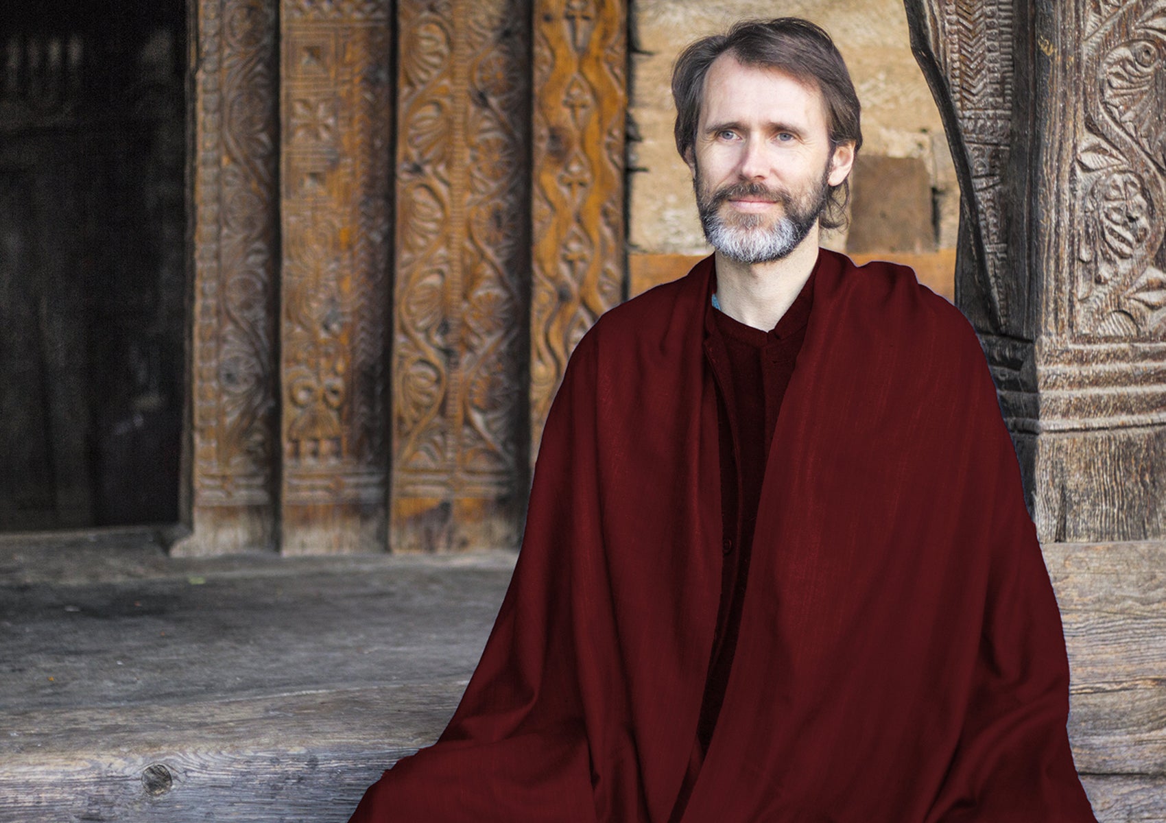 Man with a peaceful smile, wrapped in a burgundy shawl, meditating on steps of an ancient stone temple, radiating tranquility.