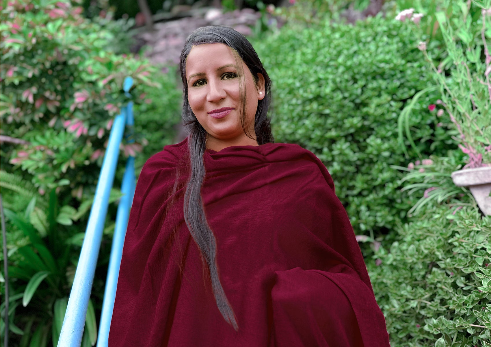 Woman wrapped in a maroon Buddhist shawl, standing outdoors with vibrant green foliage in the background, radiating tranquility and elegance.