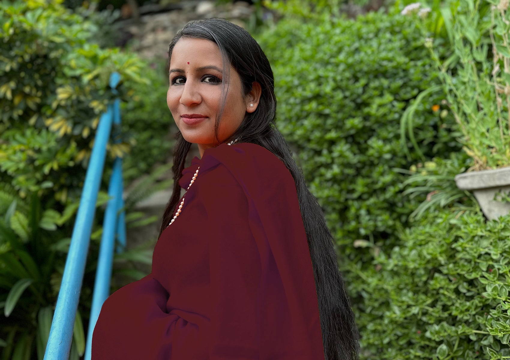 Woman draped in a maroon Buddhist shawl, smiling softly while seated on outdoor steps, surrounded by lush greenery.