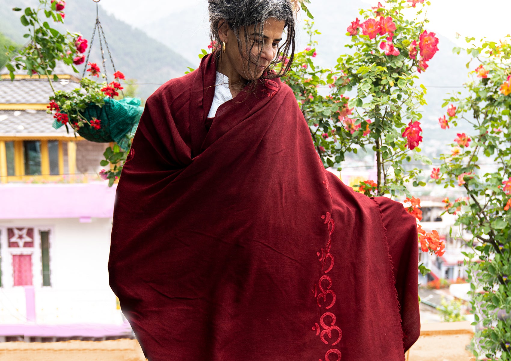 A woman wrapped in a red shawl with Om symbols, standing near vibrant flowers with mountains in the background.