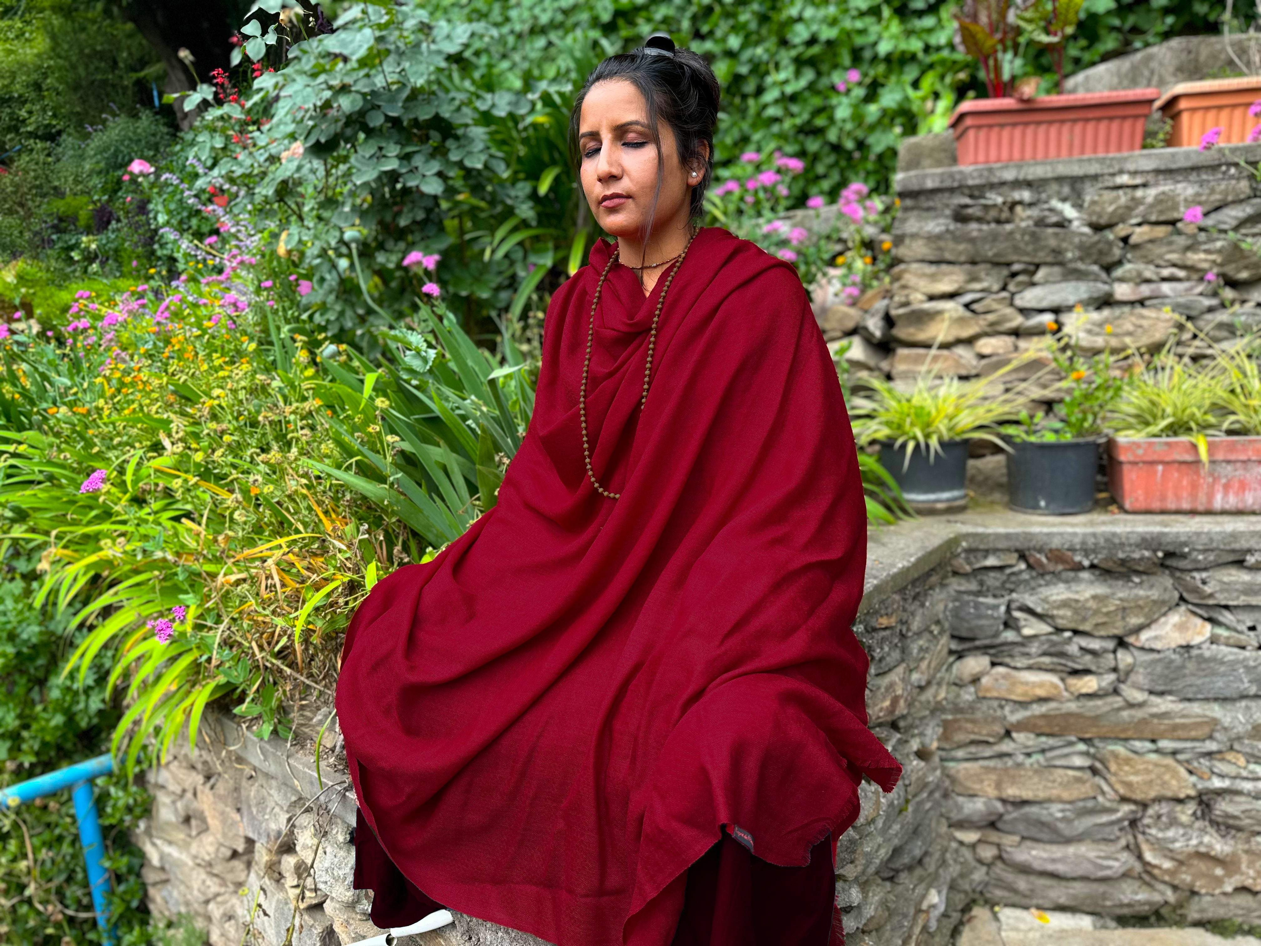 A woman seated in a red wool shawl, surrounded by green plants and blooming flowers in a garden.