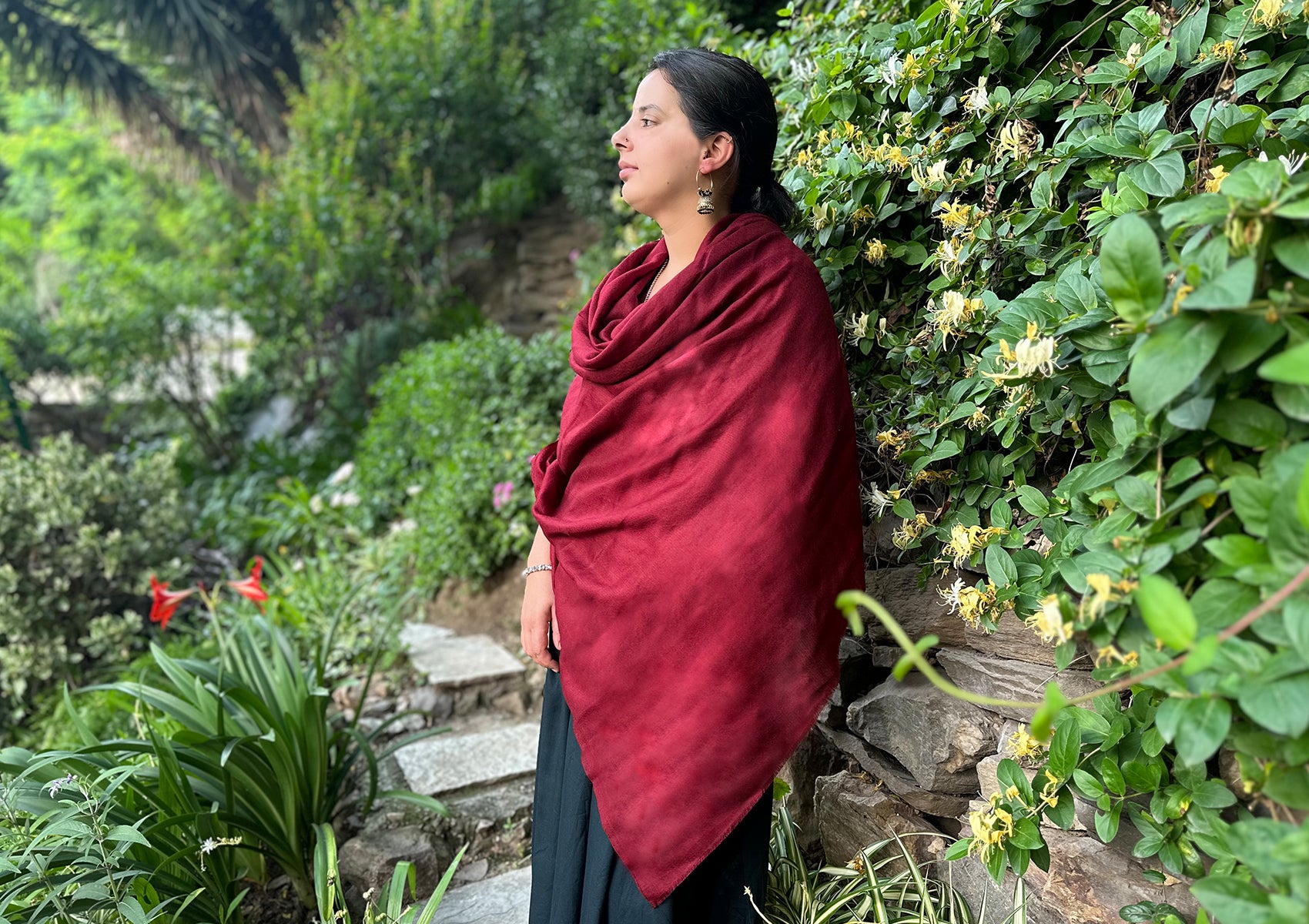 A woman wrapped in a deep red pashmina scarf standing amidst lush green garden surroundings, looking contemplatively into the distance.