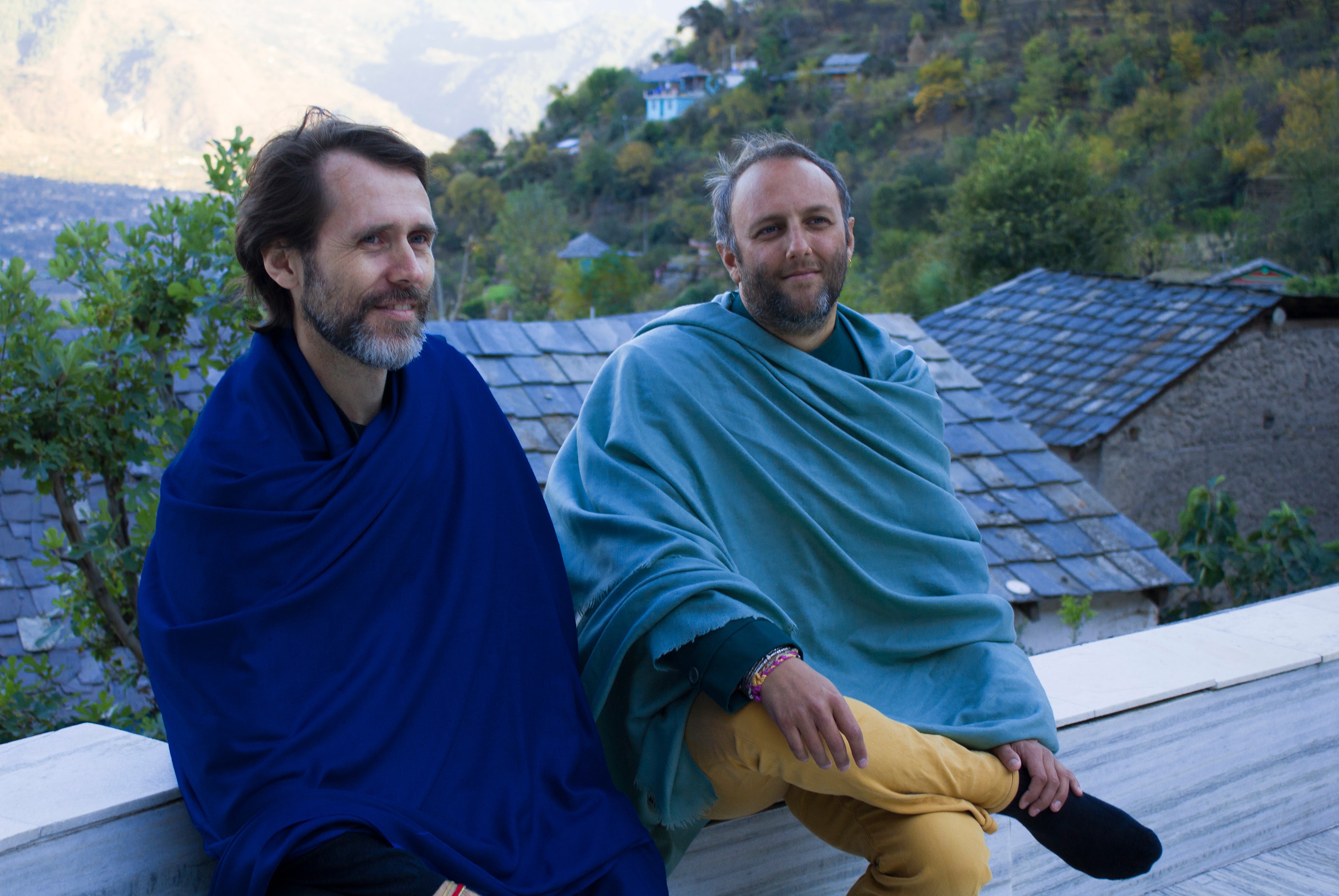 Two men seated outdoors wearing meditation shawls, one in deep blue and the other in light green, with a serene hilly backdrop.