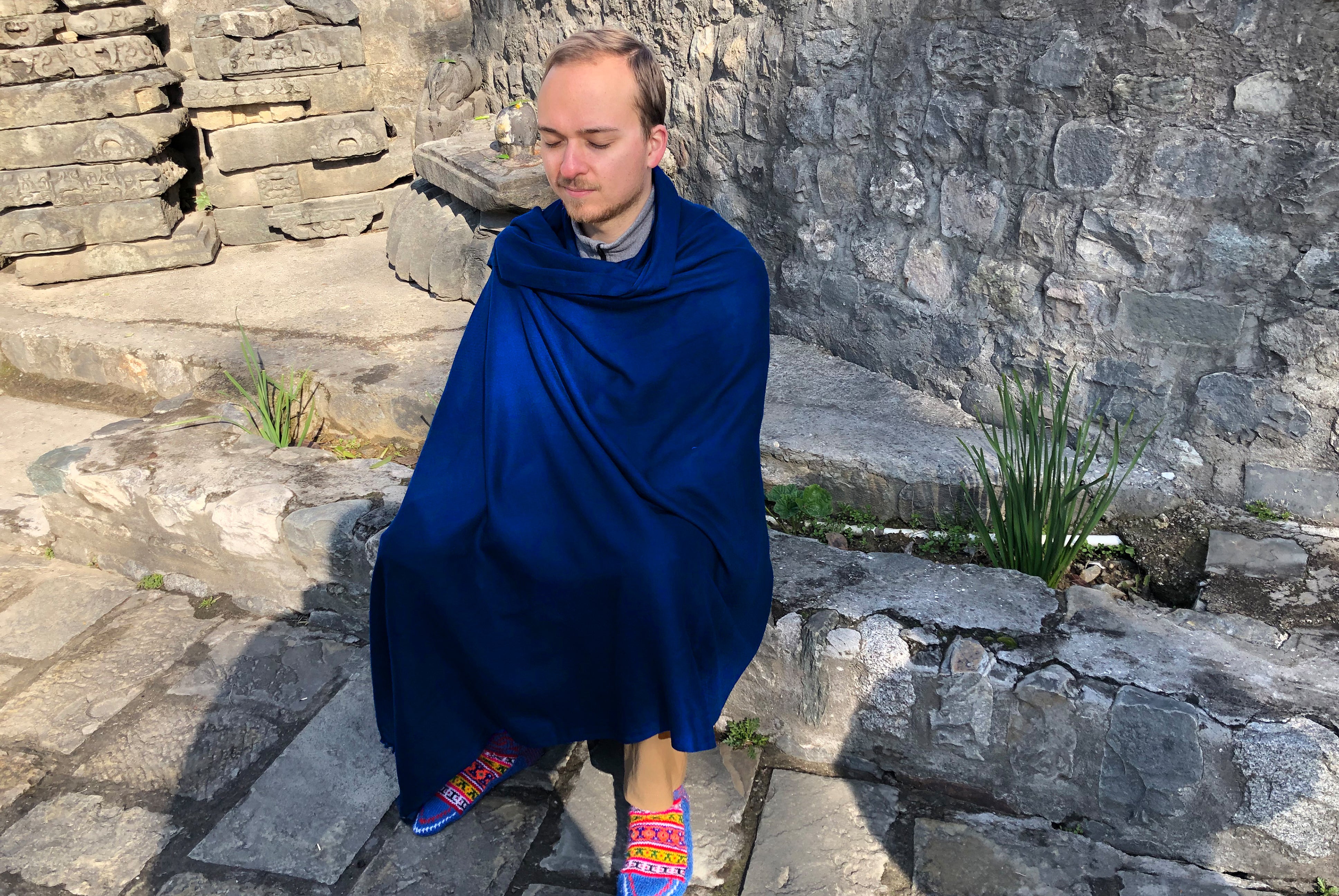 Man meditating in a royal blue shawl, seated on stone steps, surrounded by ancient architecture.