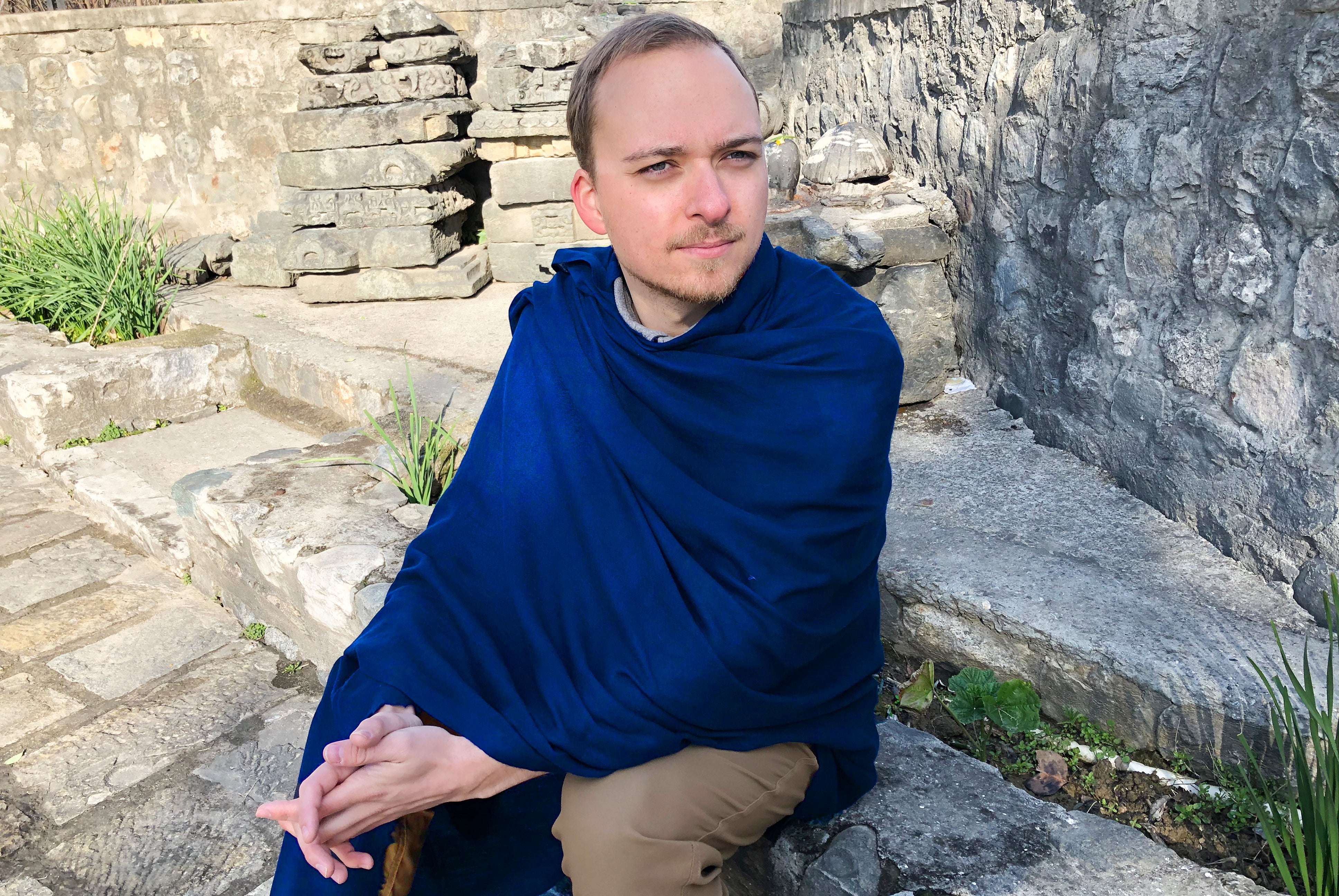 Man seated outdoors wearing a royal blue meditation shawl, exuding tranquility in a serene stone courtyard setting.