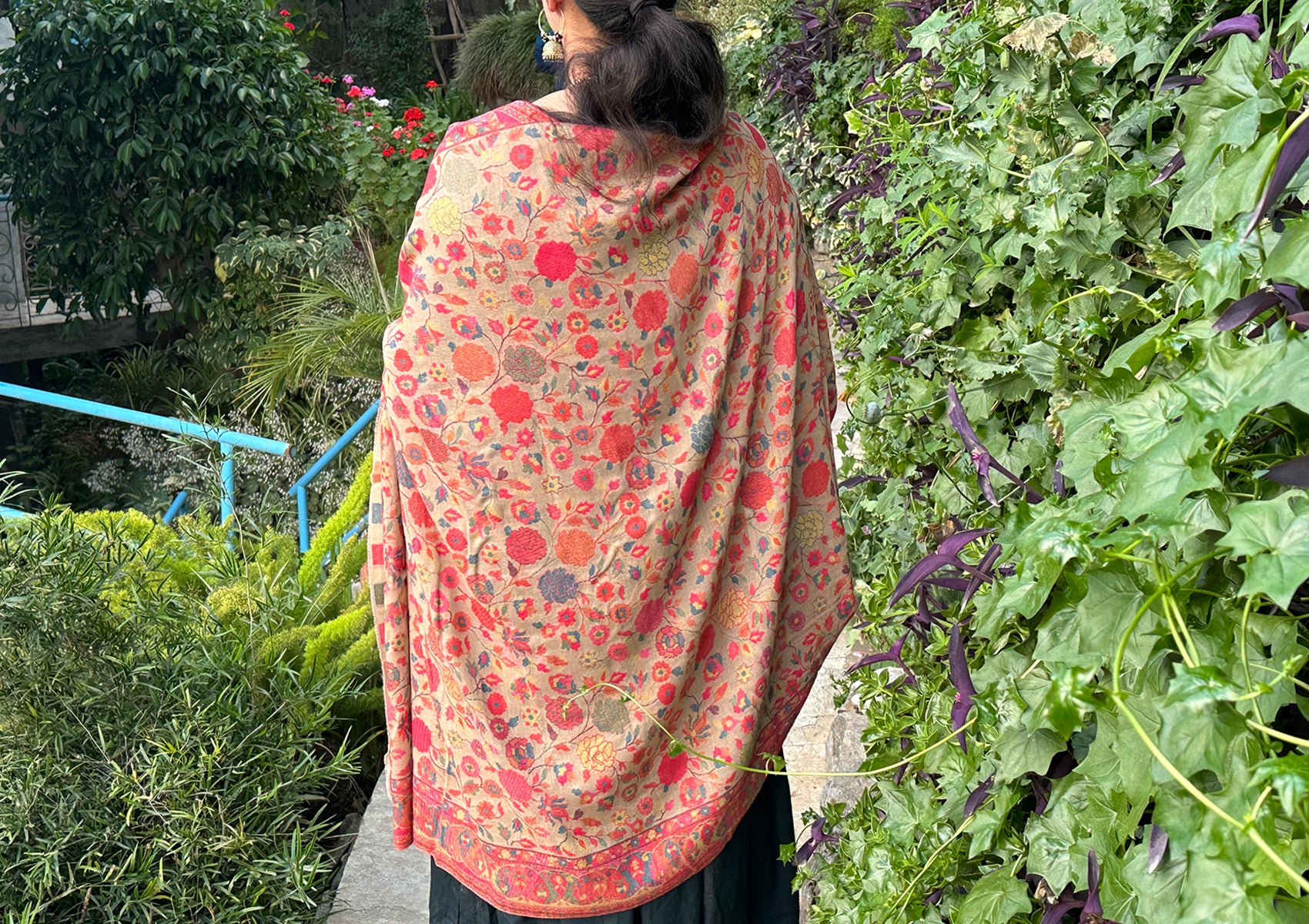 Back view of a person wearing a beige floral Mohani Kani shawl, showcasing intricate patterns and vibrant colors against a lush green garden backdrop.