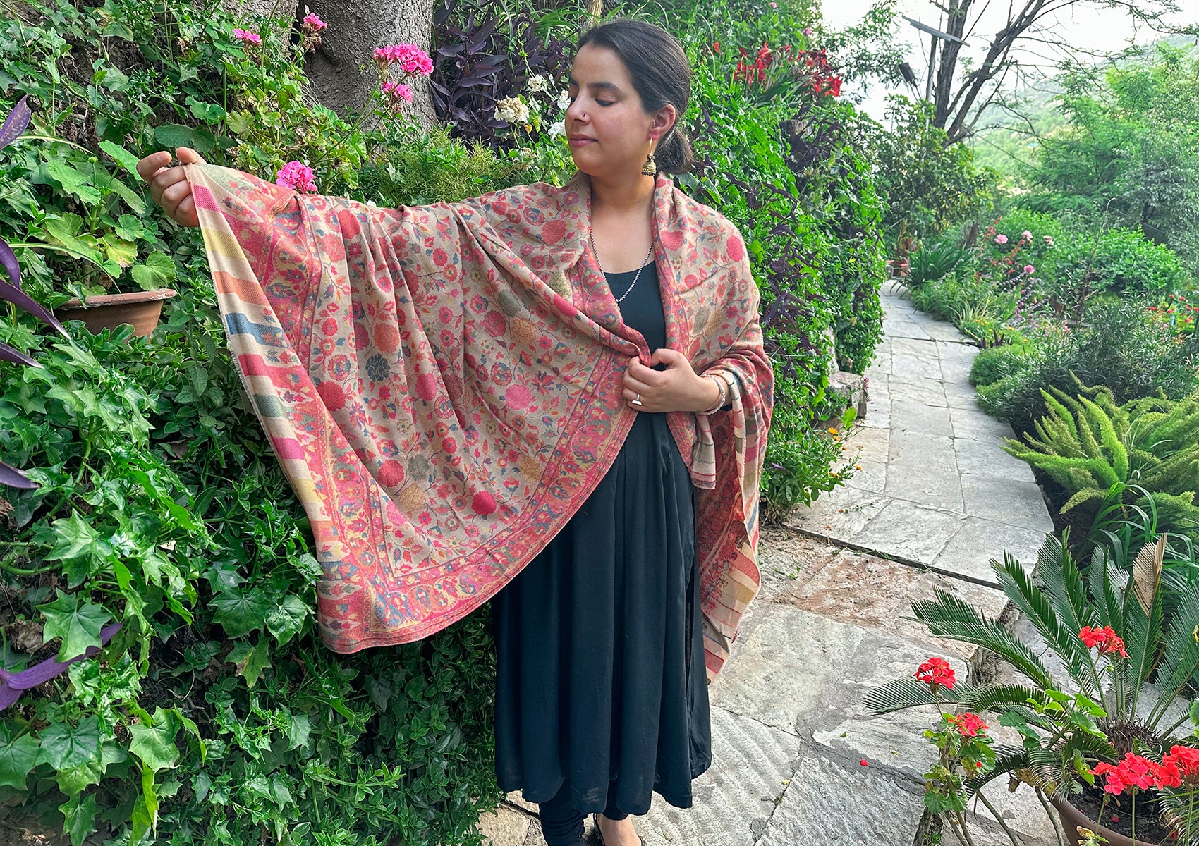 Person gracefully showcasing a beige Mohani Kani shawl with intricate floral designs, posing amidst a vibrant garden with colorful flowers and lush greenery.