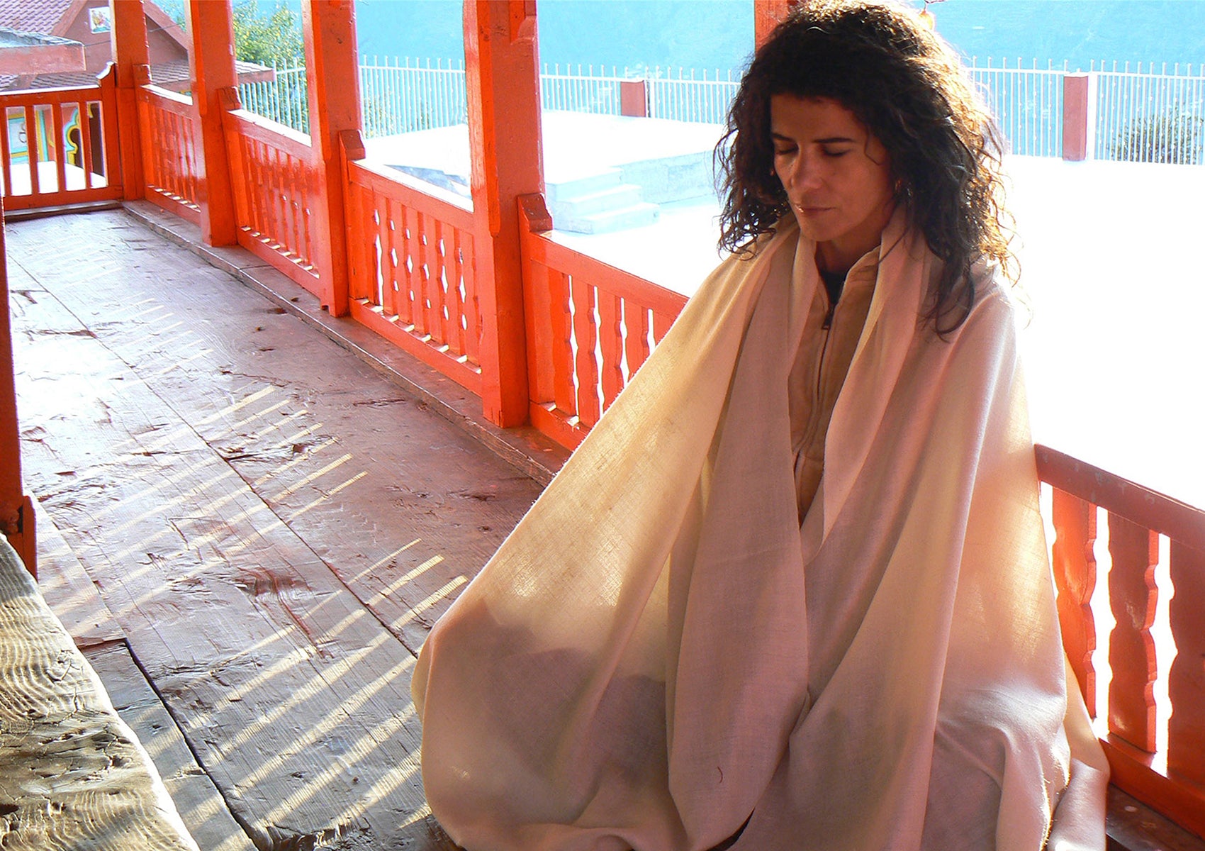 Meditating woman wearing a beige pashmina shawl, seated in a tranquil outdoor setting, under soft sunlight.