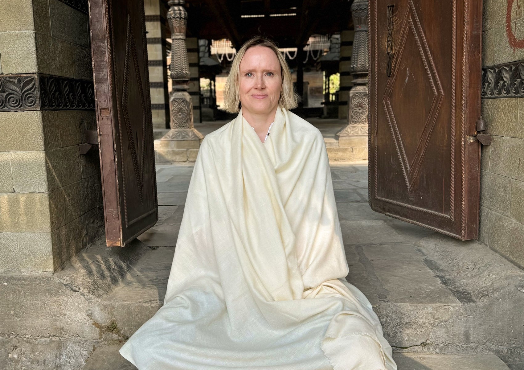 A serene portrait featuring a cream-colored Rasana Angora prayer shawl, draped elegantly around a person seated in a temple doorway. The intricate architecture highlights the spiritual ambiance.