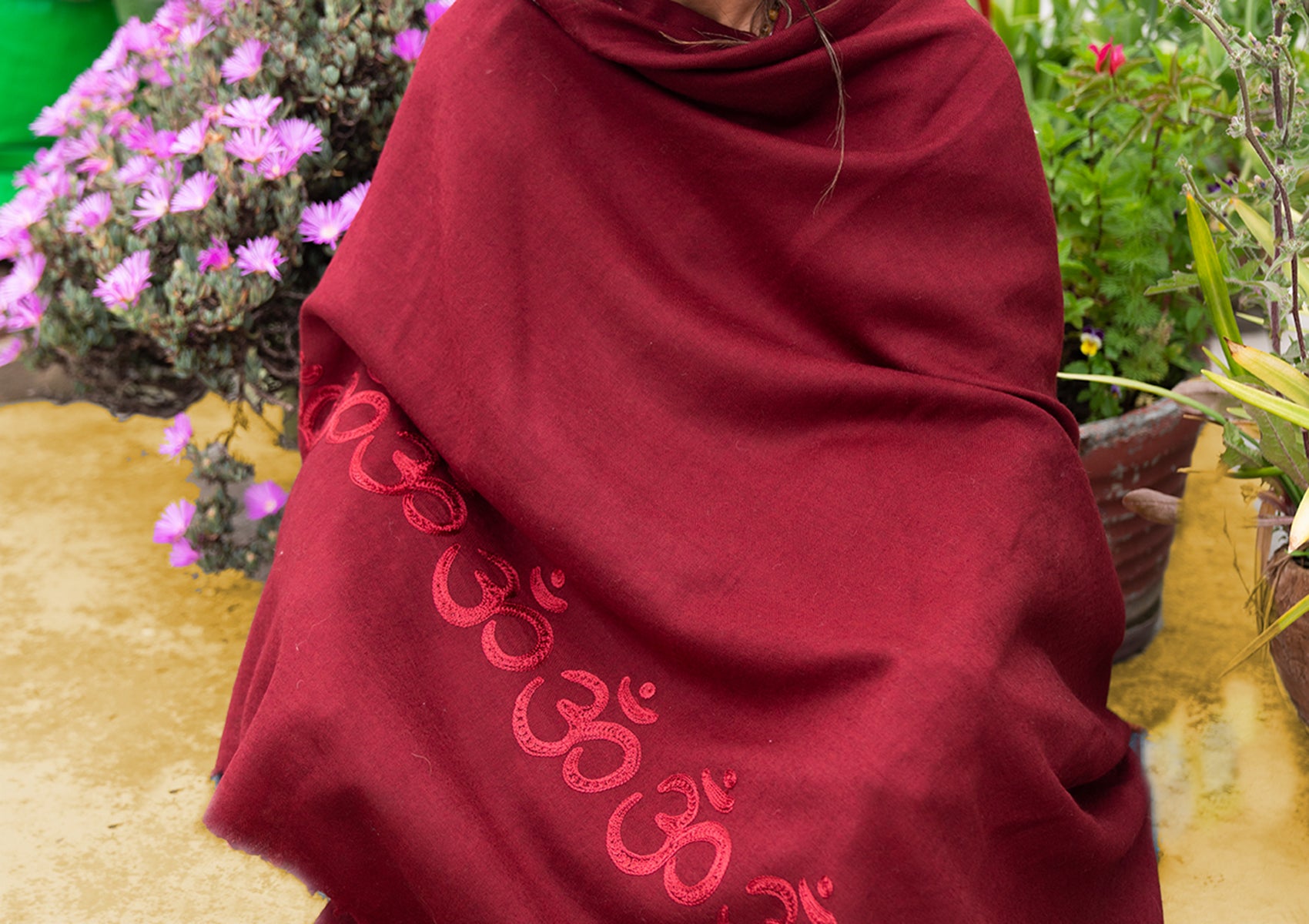 A close-up of a red shawl with embroidered Om symbols, surrounded by purple flowers, creating a serene setting.