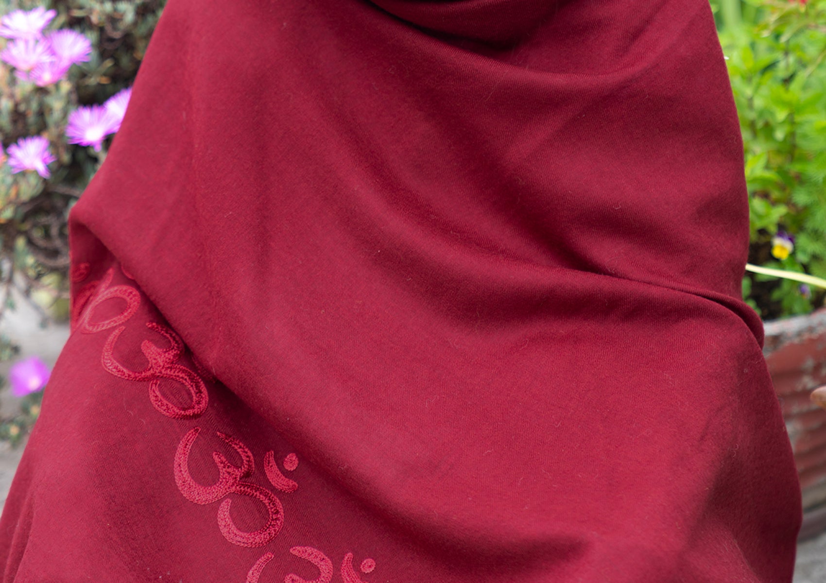 A woman seated on the ground in a red shawl with Om symbols, set against a garden backdrop, embodying calmness.