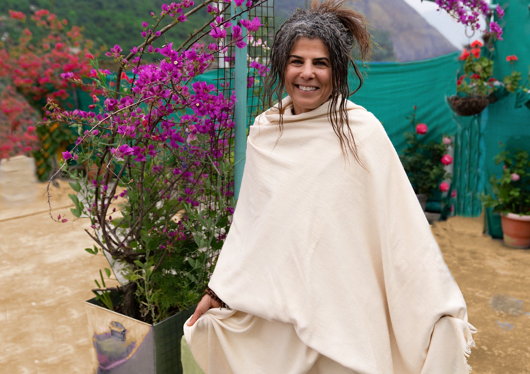 Smiling woman wrapped in a cream shawl, outdoors with mountains in the background.