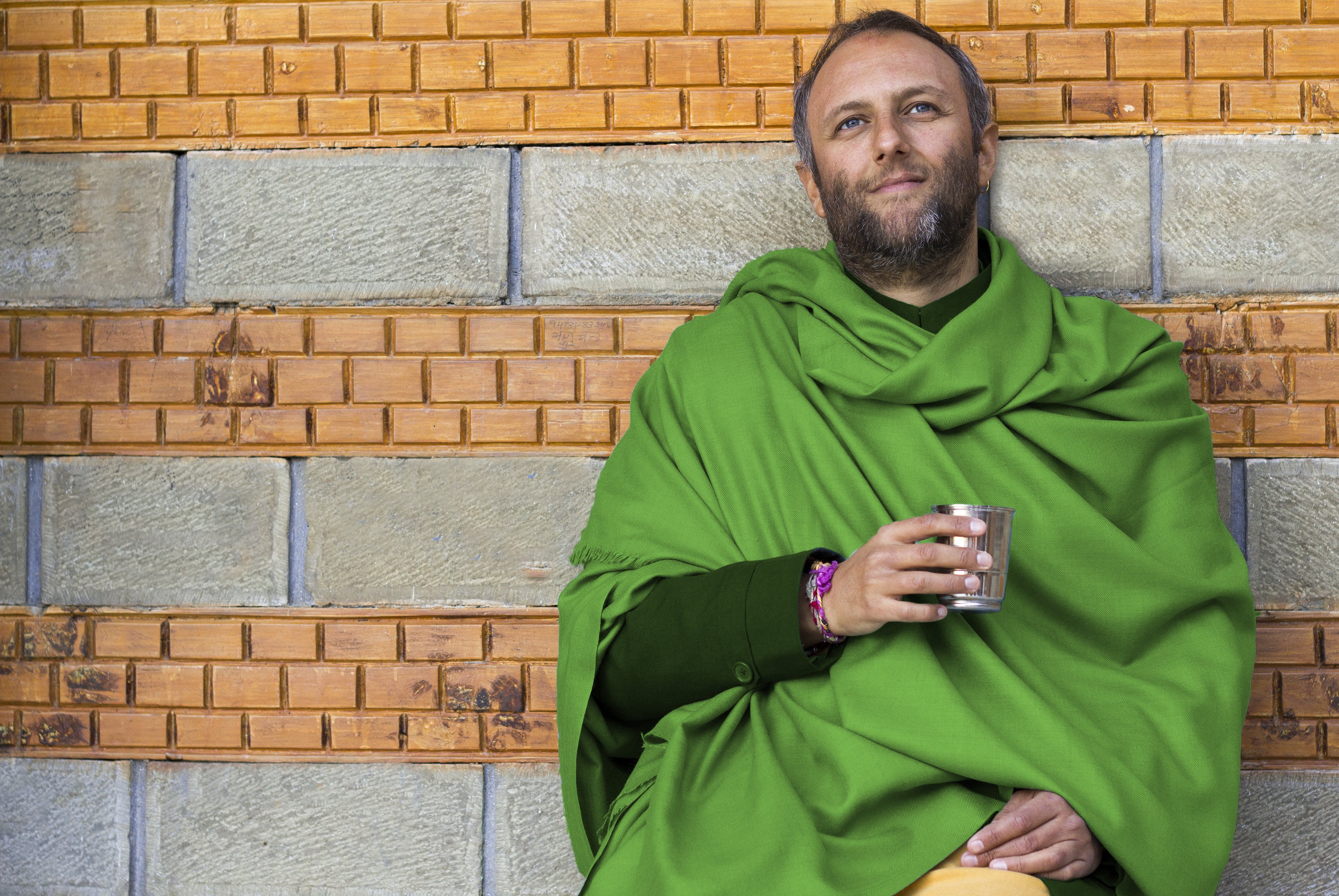 Man in a green meditation shawl seated against a textured brick and stone wall, holding a stainless steel cup