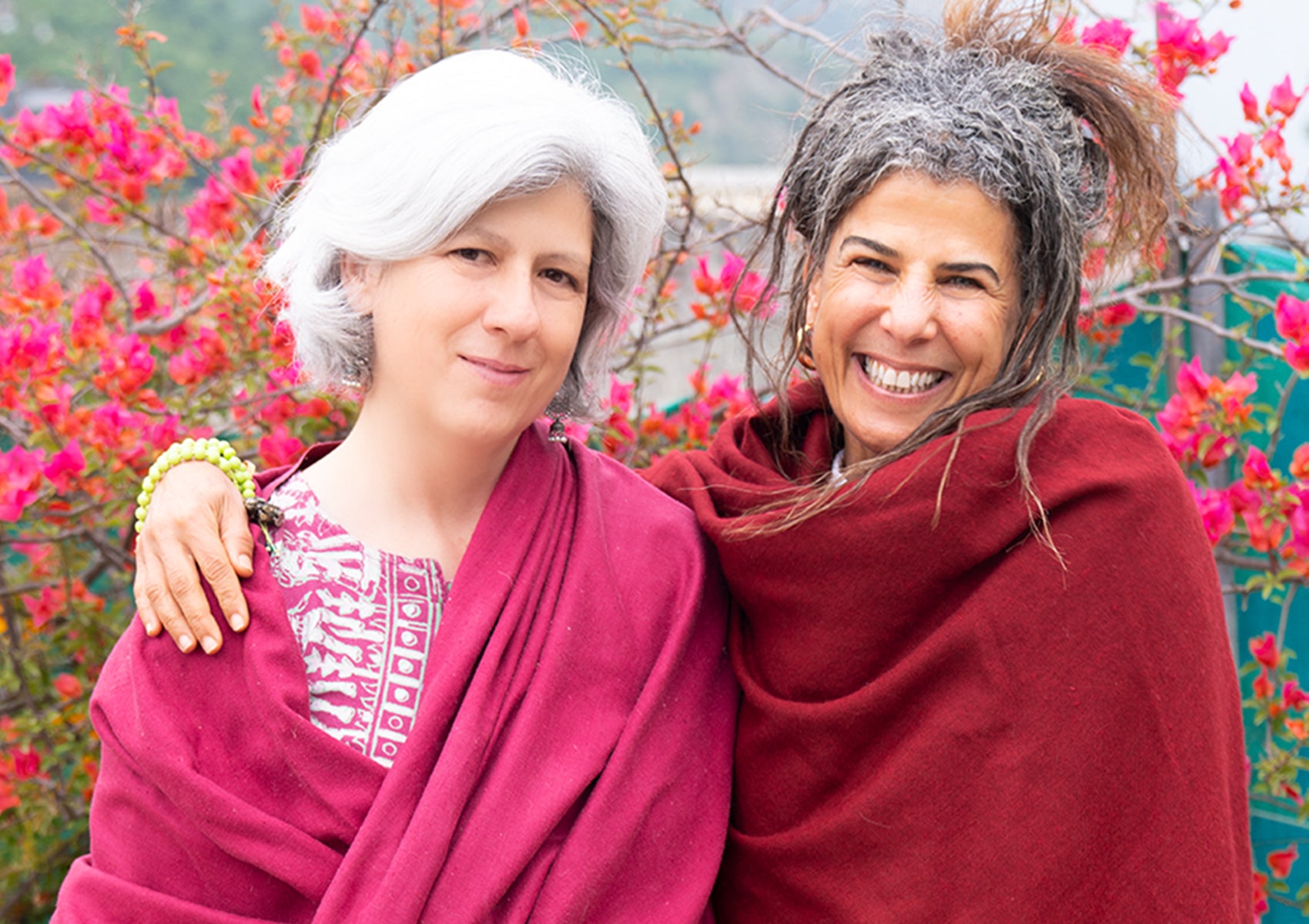 Two smiling women, one with gray hair and the other with curly hair, wrapped in burgundy and pink shawls, standing together in front of vibrant pink flowers.