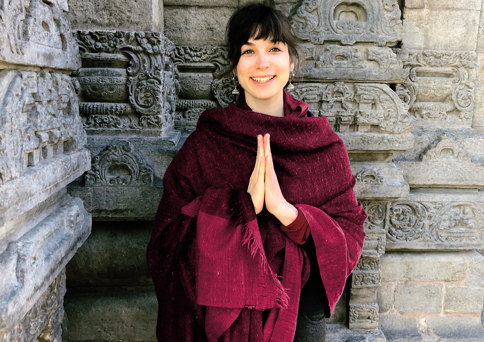 Smiling woman in a burgundy shawl with hands in a Namaste gesture, standing by a carved stone temple wall, embodying peace and cultural warmth.