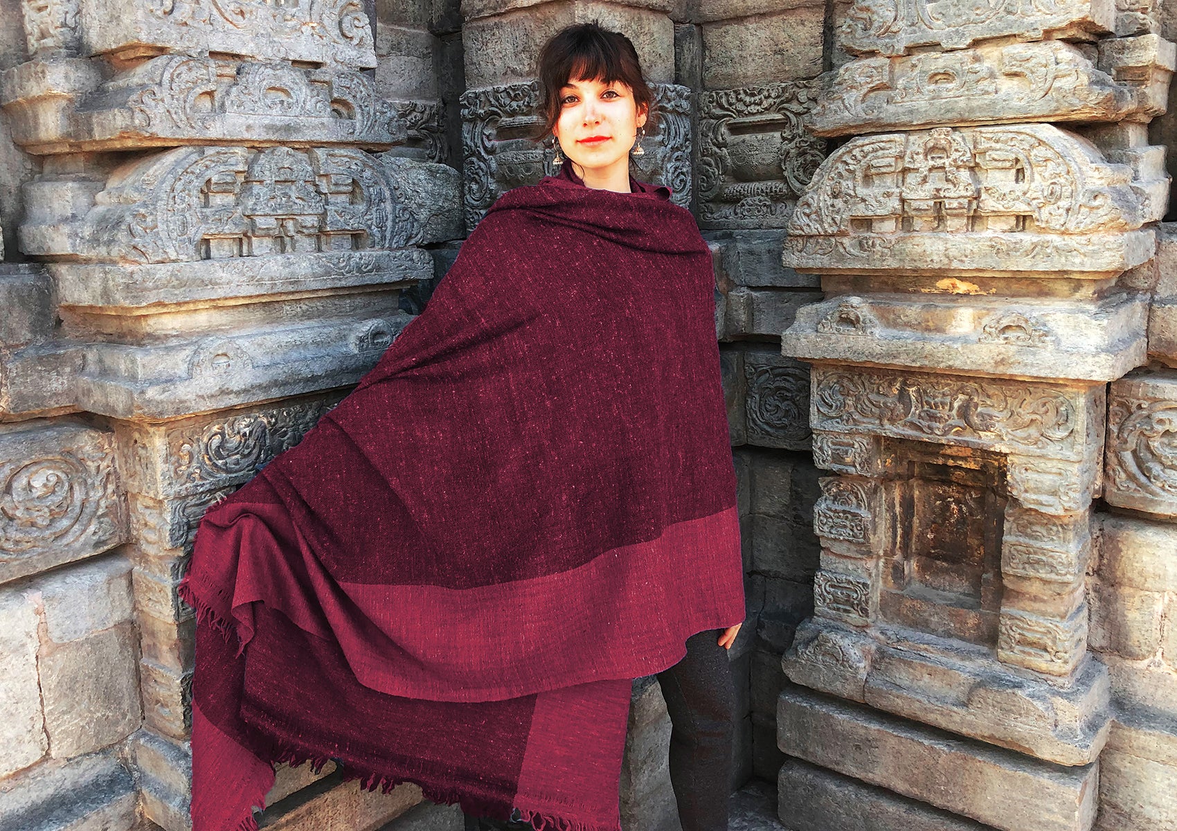 Woman wrapped in a large burgundy shawl, standing by a carved stone temple wall, displaying a blend of traditional elegance and serene style.