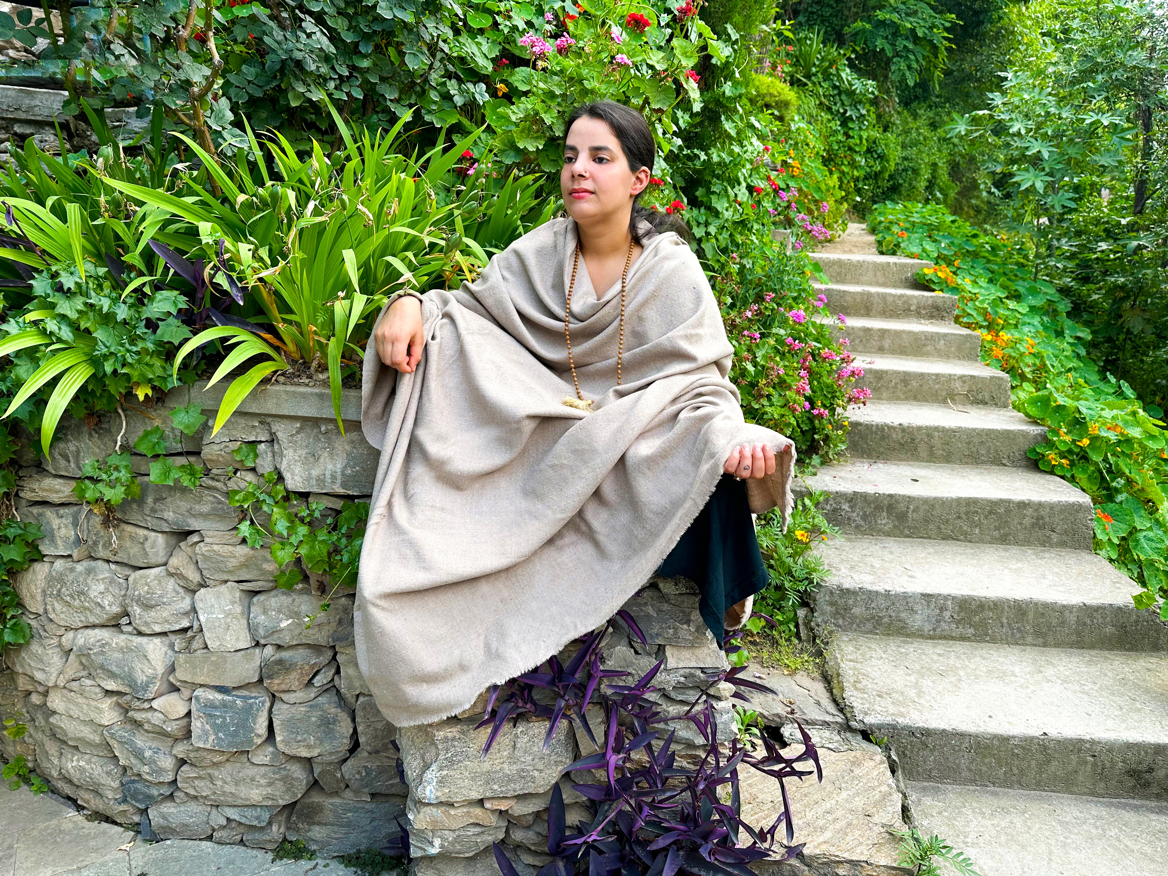 Woman seated outdoors in a lush garden, draped in a beige meditation shawl, surrounded by green plants and colorful flowers with a stone pathway in the background