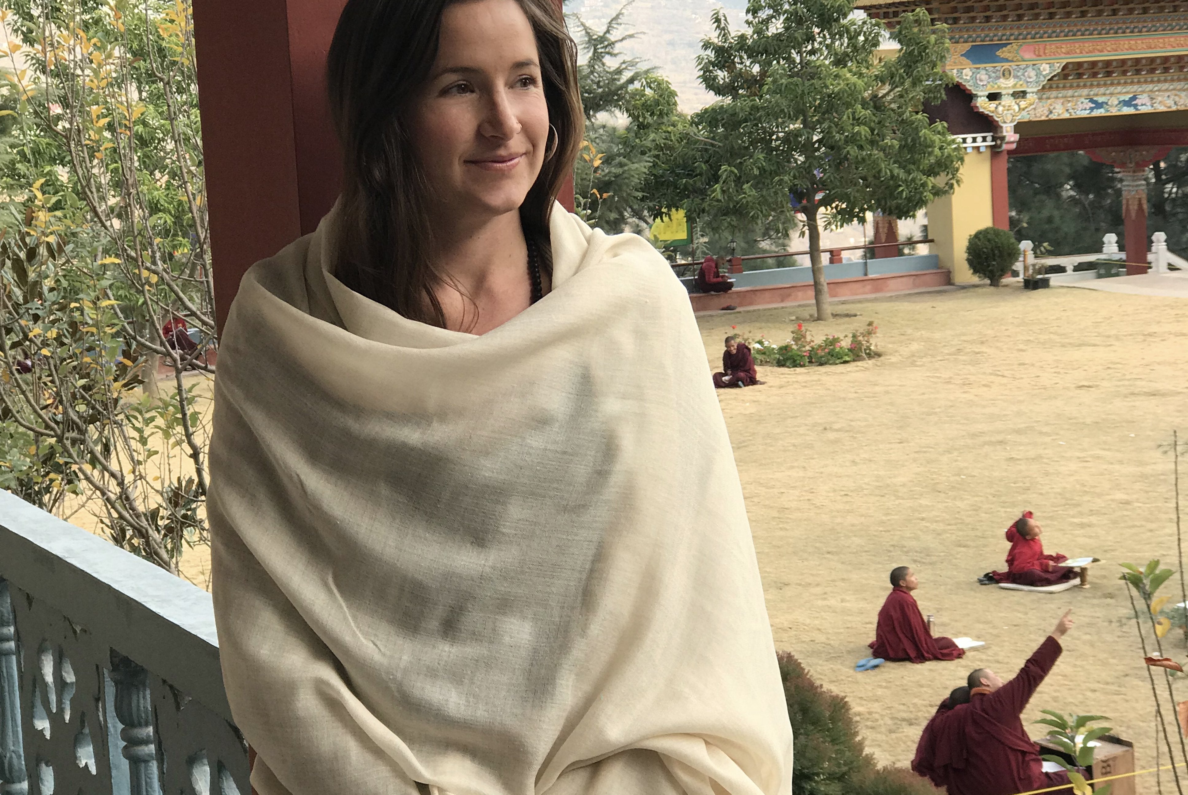 A woman wrapped in a beige shawl, standing on a balcony with monks seated on the ground in the background at a monastery courtyard