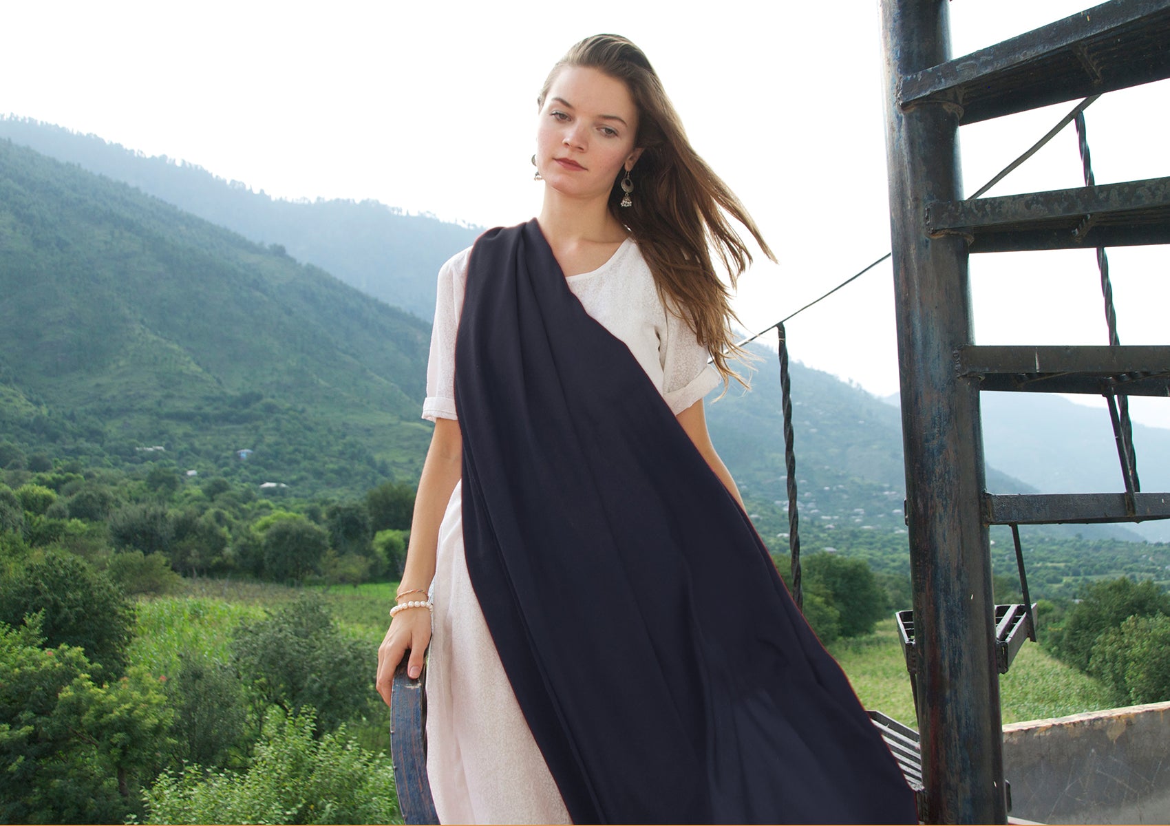 A woman wearing a navy blue shawl stands in front of a scenic mountain backdrop, with the wind gently blowing her hair, creating a peaceful and serene atmosphere.