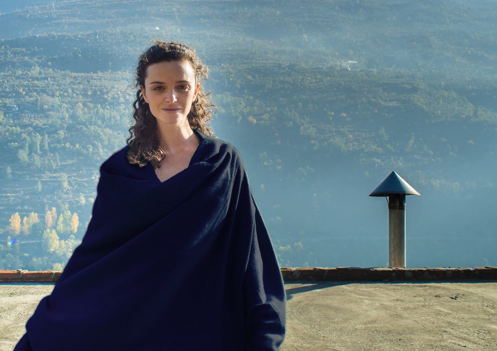 A woman wrapped in a deep navy pashmina shawl stands in front of a scenic mountain landscape under soft sunlight, radiating calm and tranquility