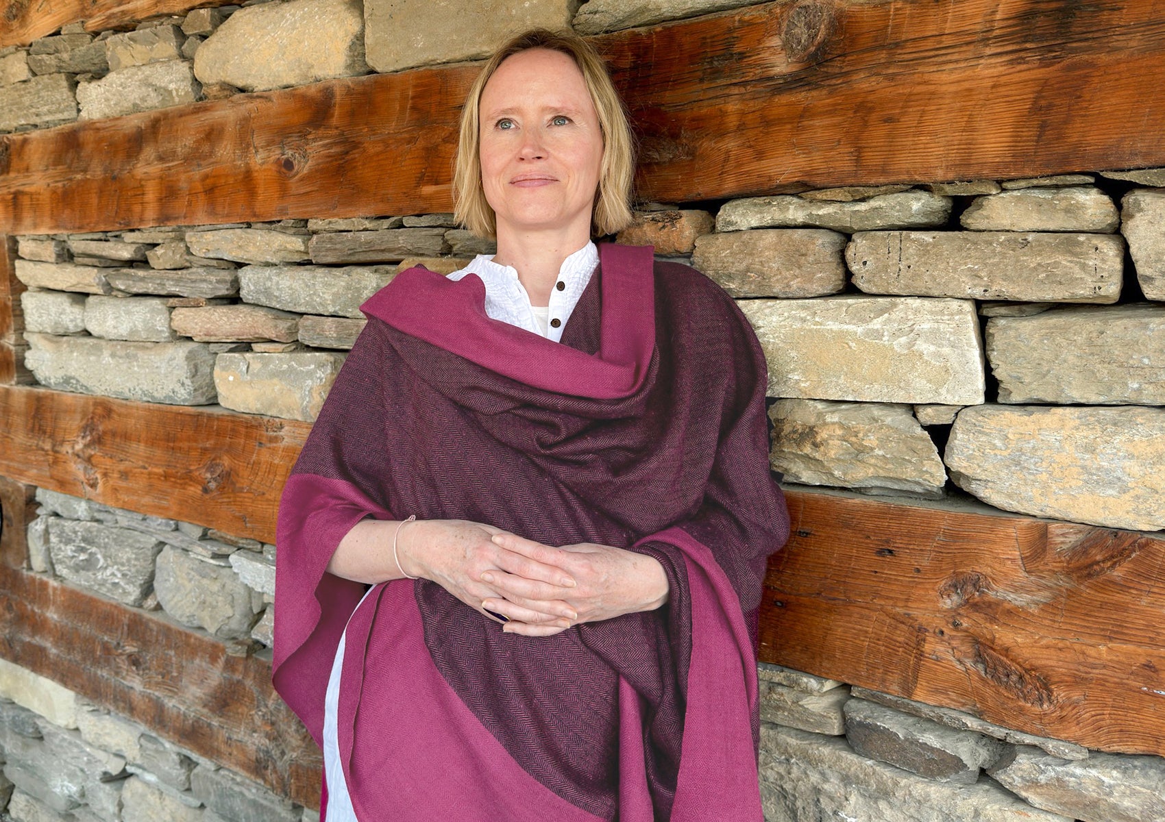 Woman wearing a magenta shawl, standing against a rustic stone wall with wooden accents, exuding calm and natural elegance.