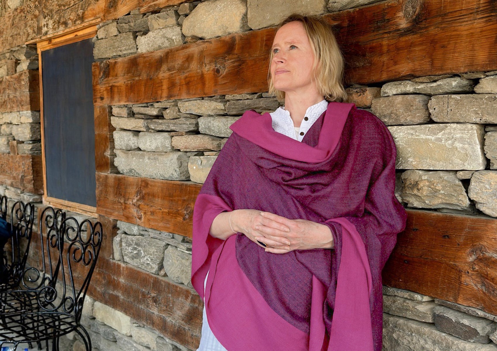 Woman in a magenta shawl leaning against a rustic stone wall, gazing thoughtfully into the distance, capturing a serene and contemplative moment.