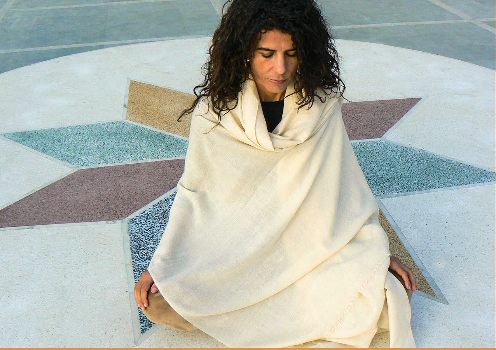 A woman wrapped in a beige pashmina shawl sitting cross-legged in meditation on a colorful geometric-patterned floor