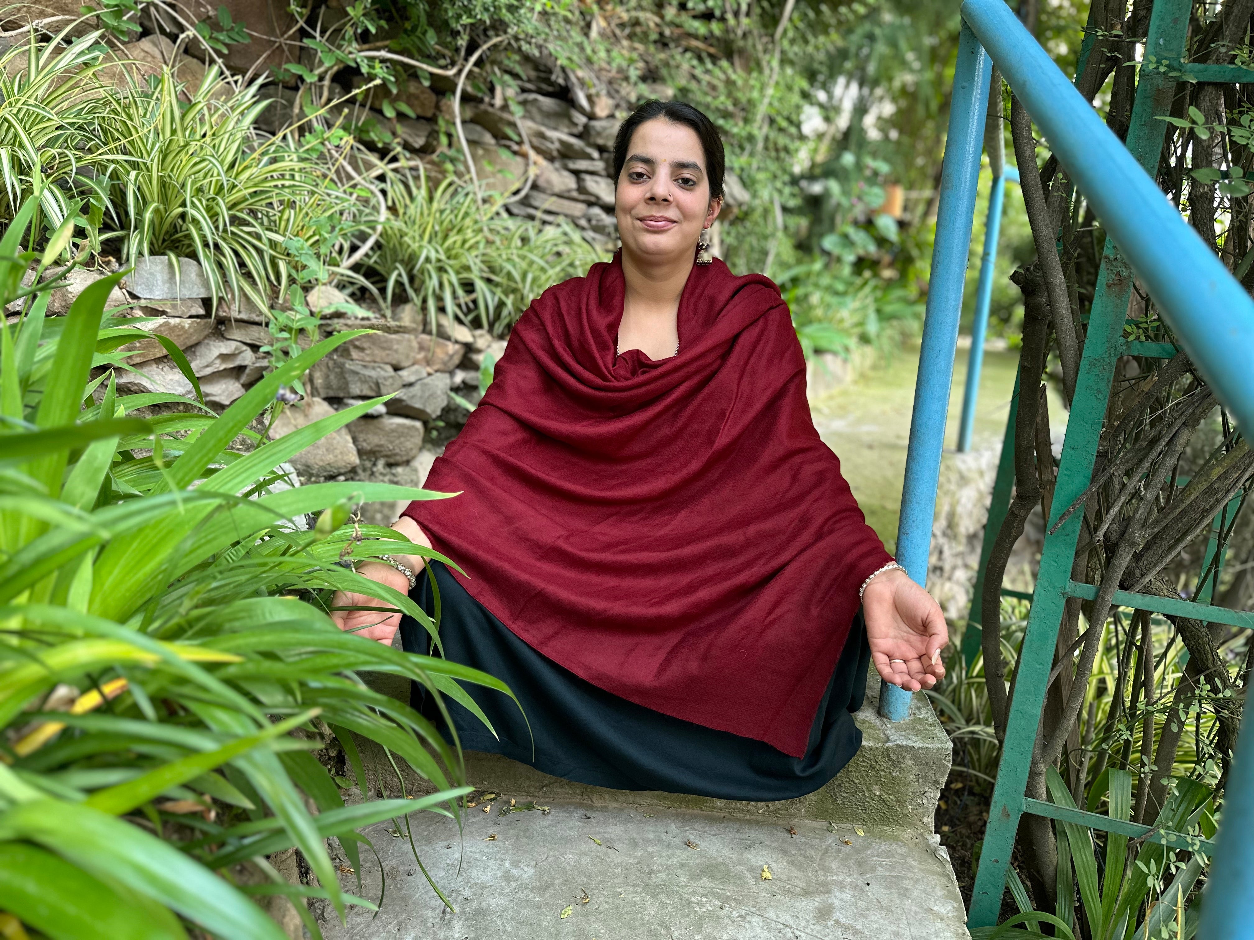 A woman wearing a red pashmina shawl seated in a meditative pose outdoors, surrounded by green plants, near a rustic stone wall and blue metal railing.