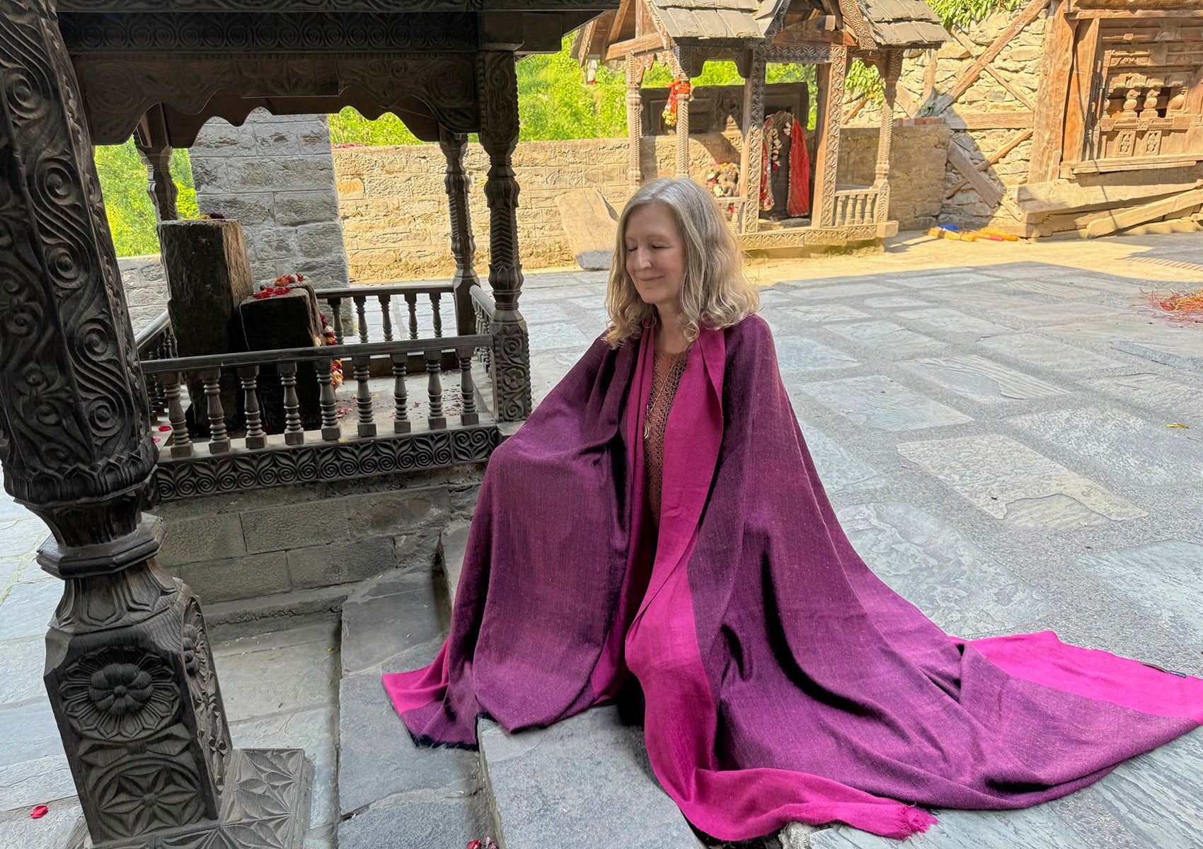 Woman wrapped in a magenta shawl, meditating peacefully at an ancient temple with intricate wood carvings, surrounded by a serene atmosphere.