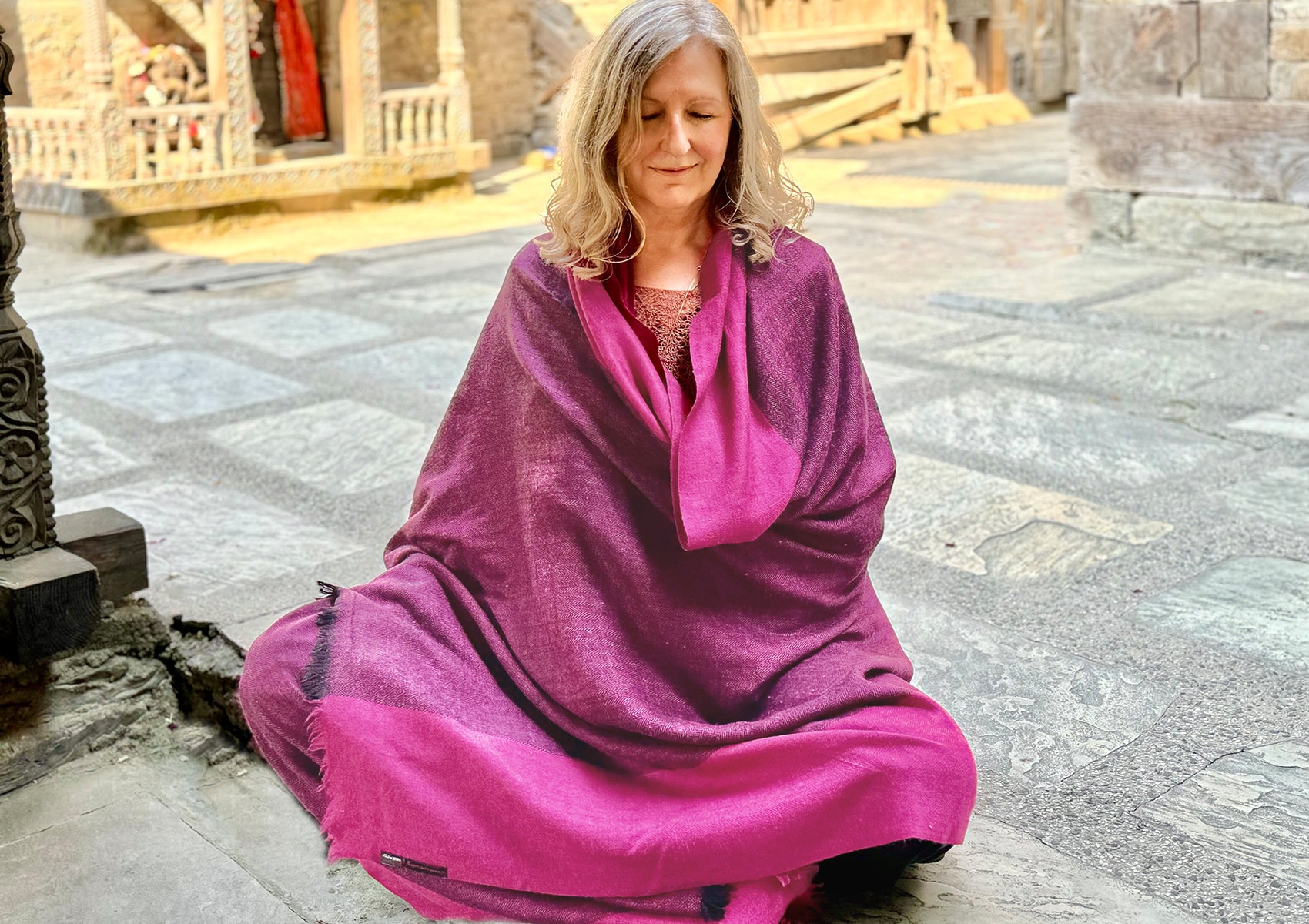 Woman meditating in a magenta shawl, seated cross-legged on stone floor with a serene temple background, exuding calmness and tranquility.