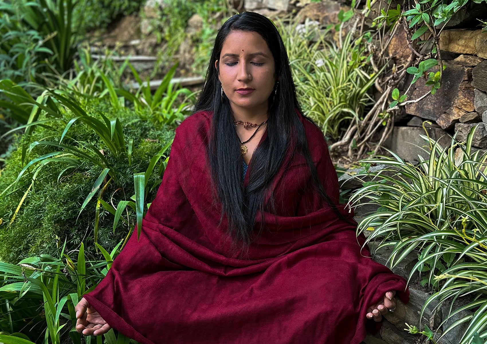 Woman meditating outdoors, wrapped in a deep red pashmina shawl, surrounded by lush greenery