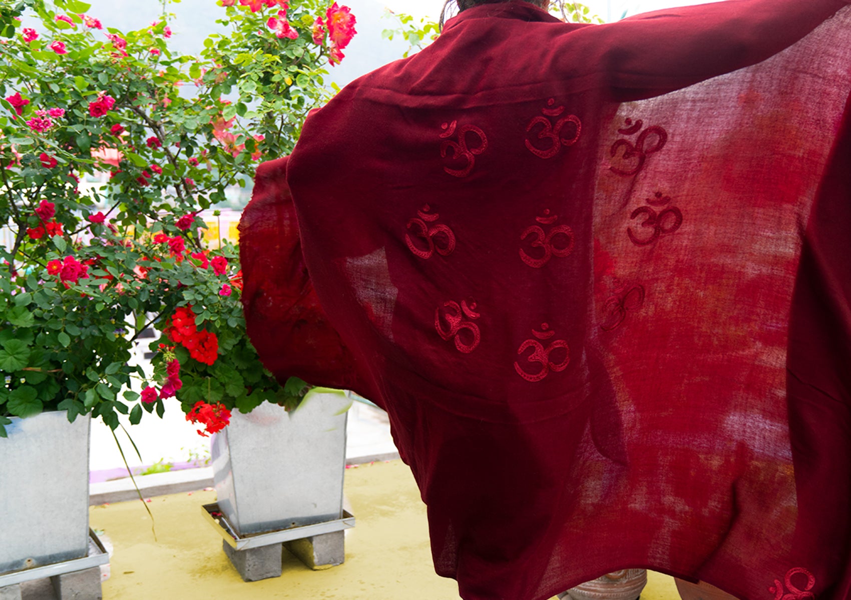 Back view of a woman with a red Om shawl, surrounded by colorful flowers, blending with nature.