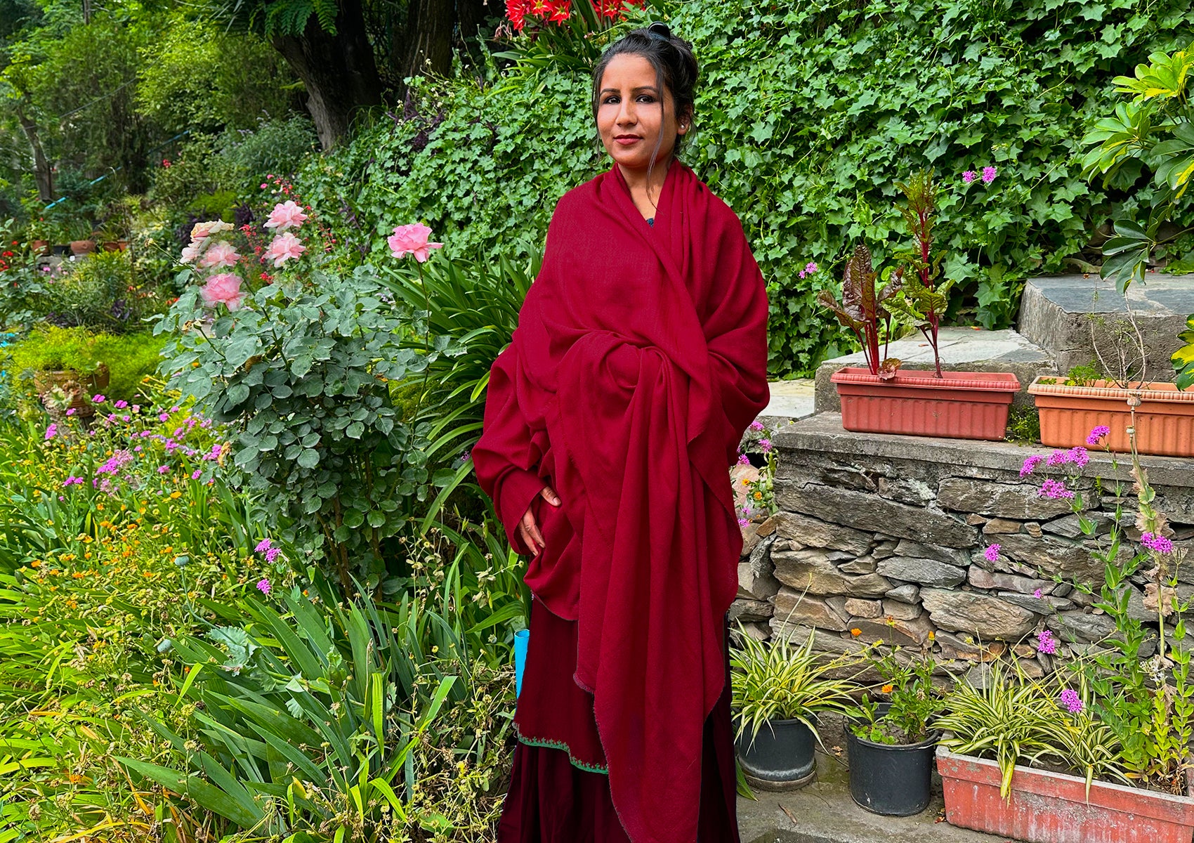 A woman standing in a lush garden wearing a red wool shawl, embracing tranquility and nature.