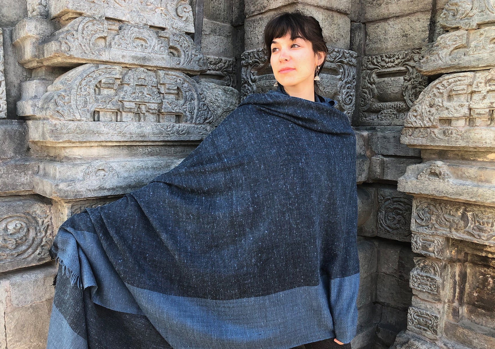 Woman wrapped in a large blue shawl, standing gracefully against a stone temple wall with intricate carvings, merging elegance with cultural heritage.